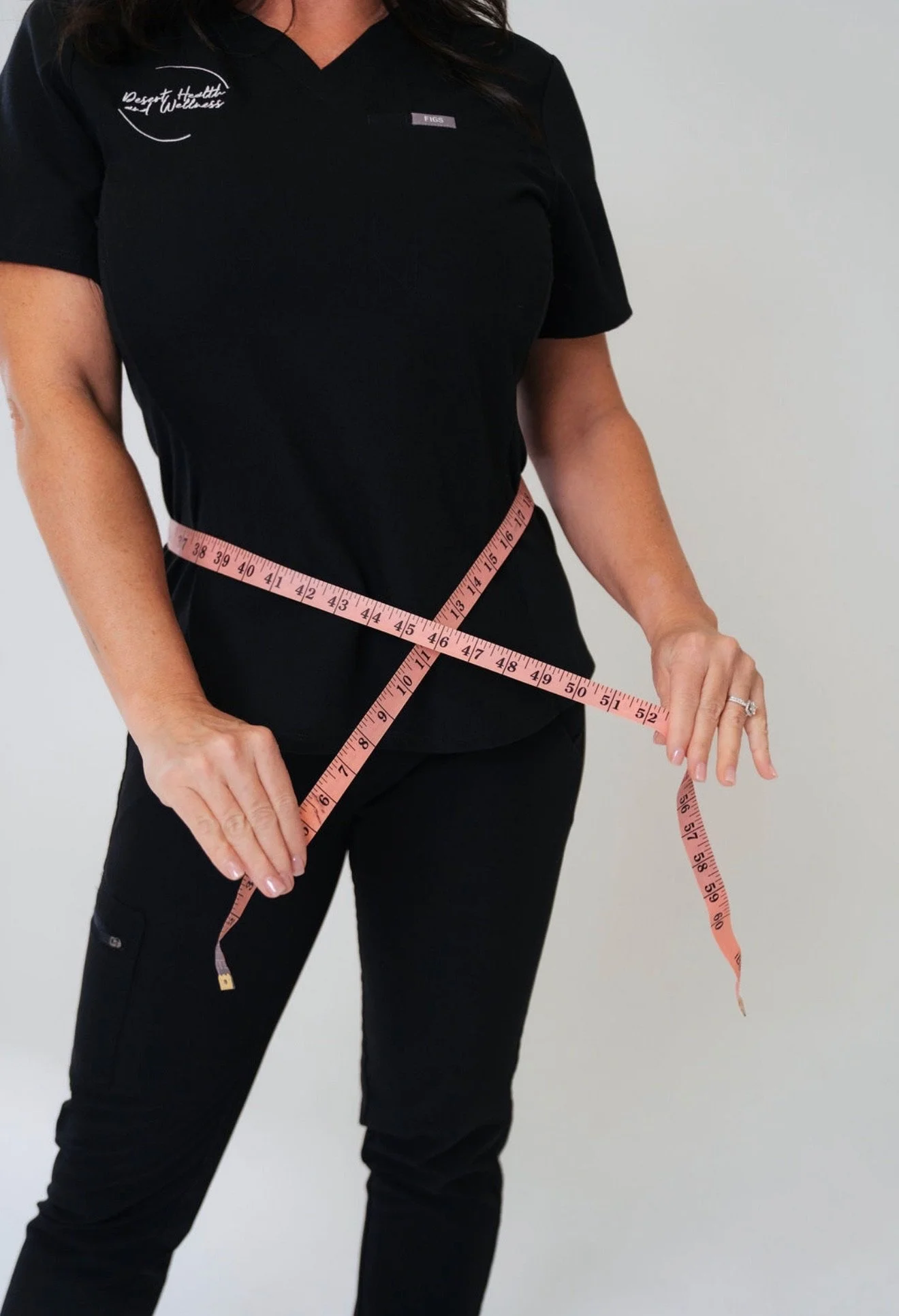 A person wearing a black scrub shirt with text on the left chest area and a small rectangular name tag, measuring their waist with a pink measuring tape. The person is also wearing black pants.