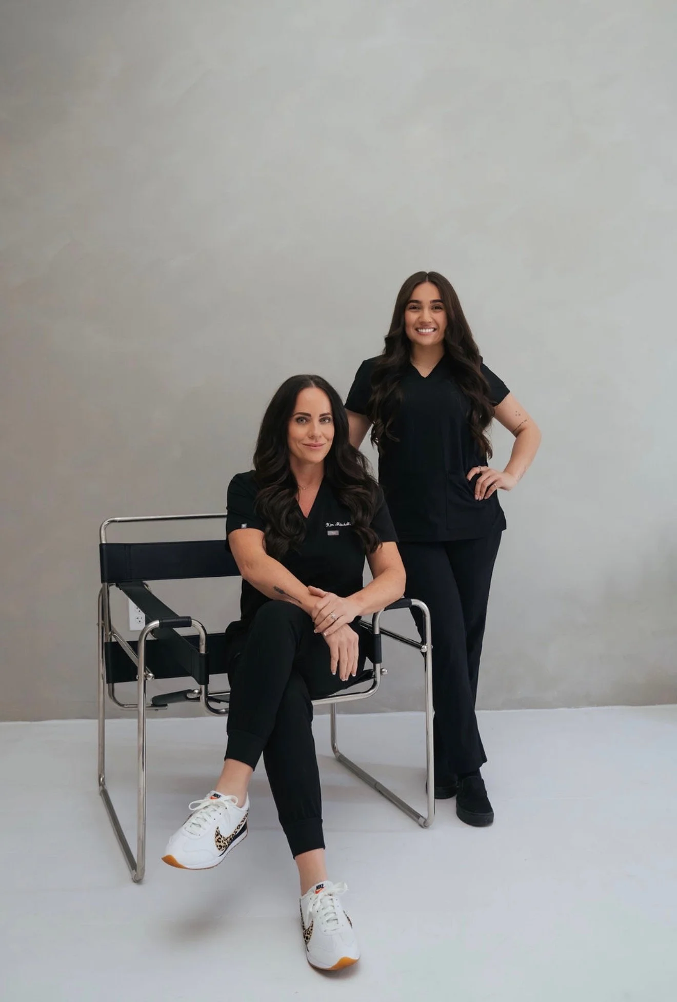 Two women in black medical scrubs pose for a photo against a plain gray wall. One woman is sitting on a chrome chair with black seat and back, while the other stands behind her with one hand on her waist. The seated woman has long, wavy dark hair and is wearing white sneakers with leopard print accents. The standing woman has long, wavy hair and is smiling with her hand on her hip.