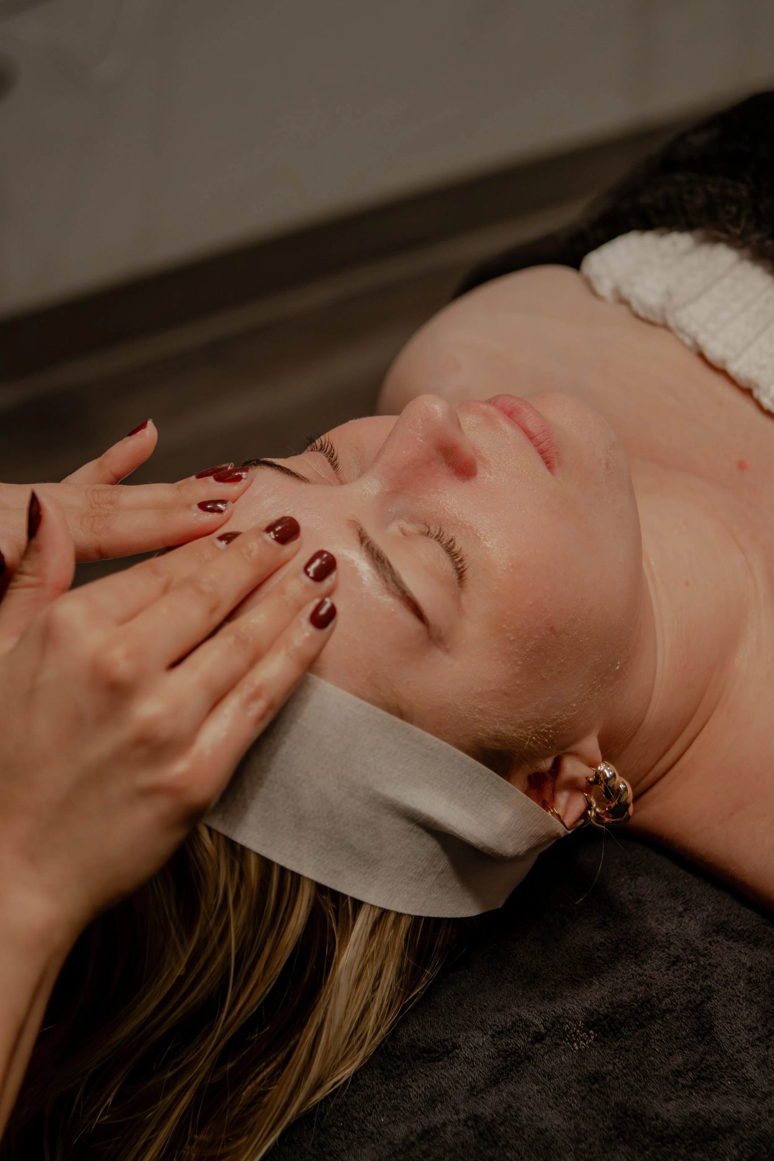 A woman lying on a massage table receiving a facial treatment, with her eyes closed and her face being gently massaged by a professional's hands.