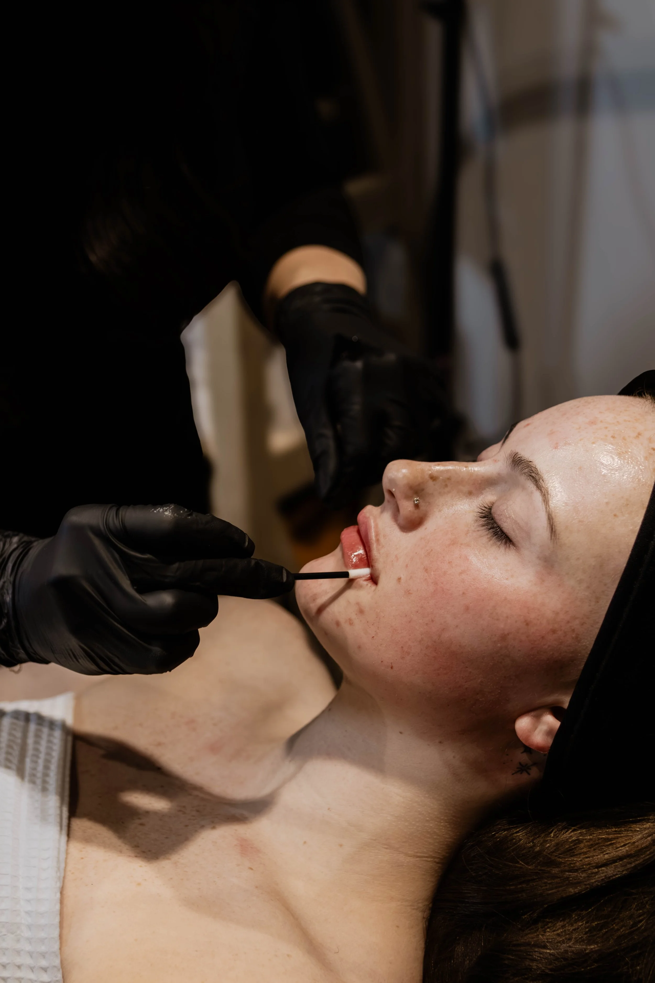 A person lying down with their eyes closed, receiving a cosmetic or skincare treatment on their lips from a professional wearing black gloves.
