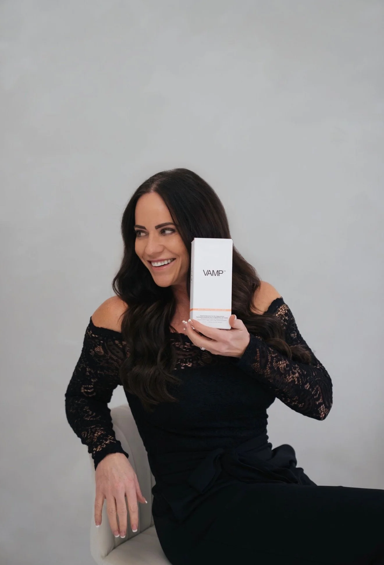 A woman with long dark wavy hair, wearing a black lace top, is smiling while holding a white box labeled 'VAMP' near her face. She is sitting on a white chair against a plain gray wall.