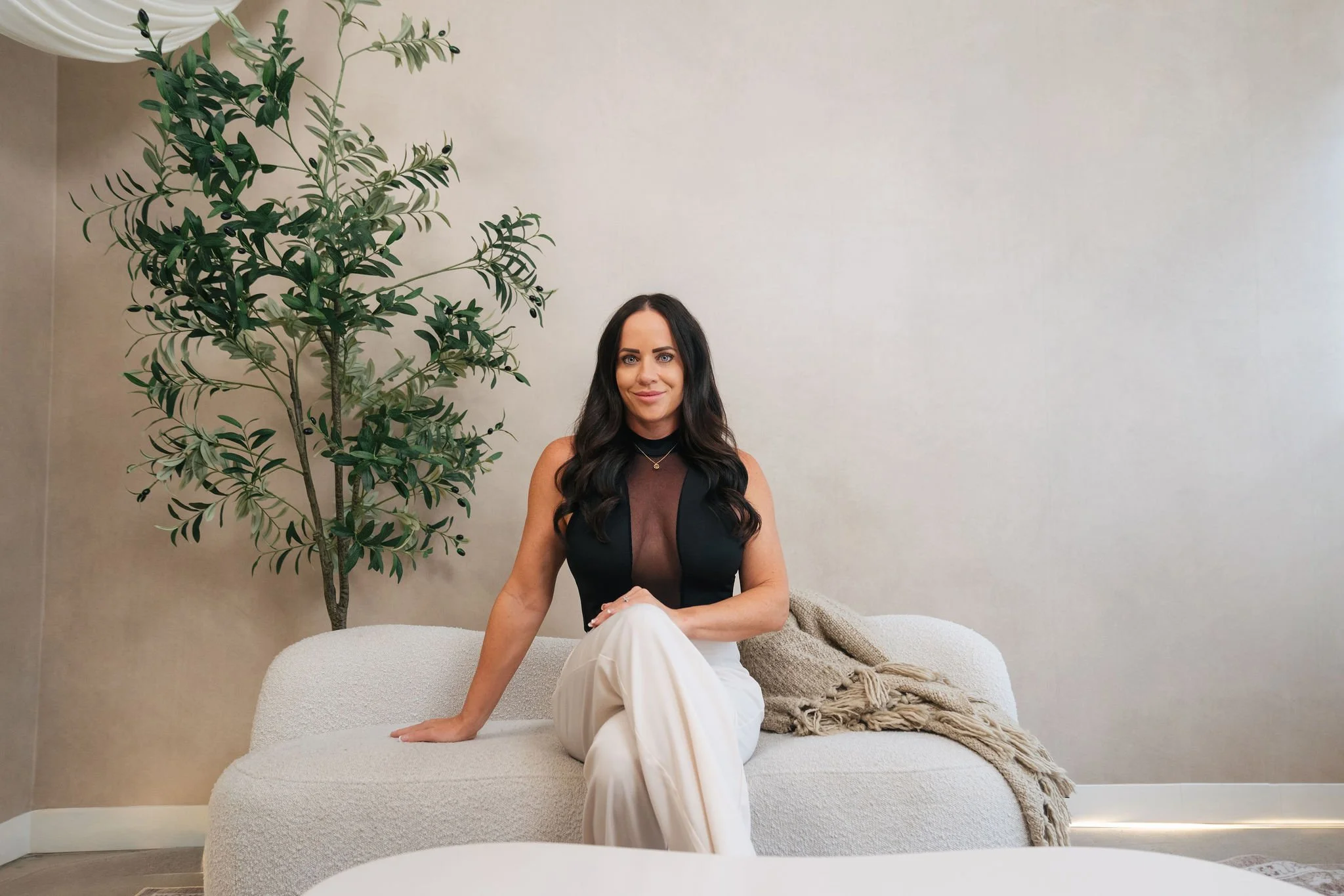 A woman with long dark hair sitting on a light-colored sofa, with a large green potted plant next to her, in a room with plain beige walls and a throw blanket.