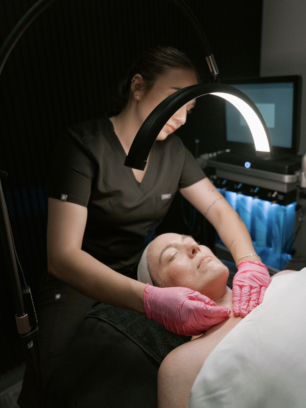 A woman performing a facial treatment on a client lying down with eyes closed in a spa or skincare clinic.