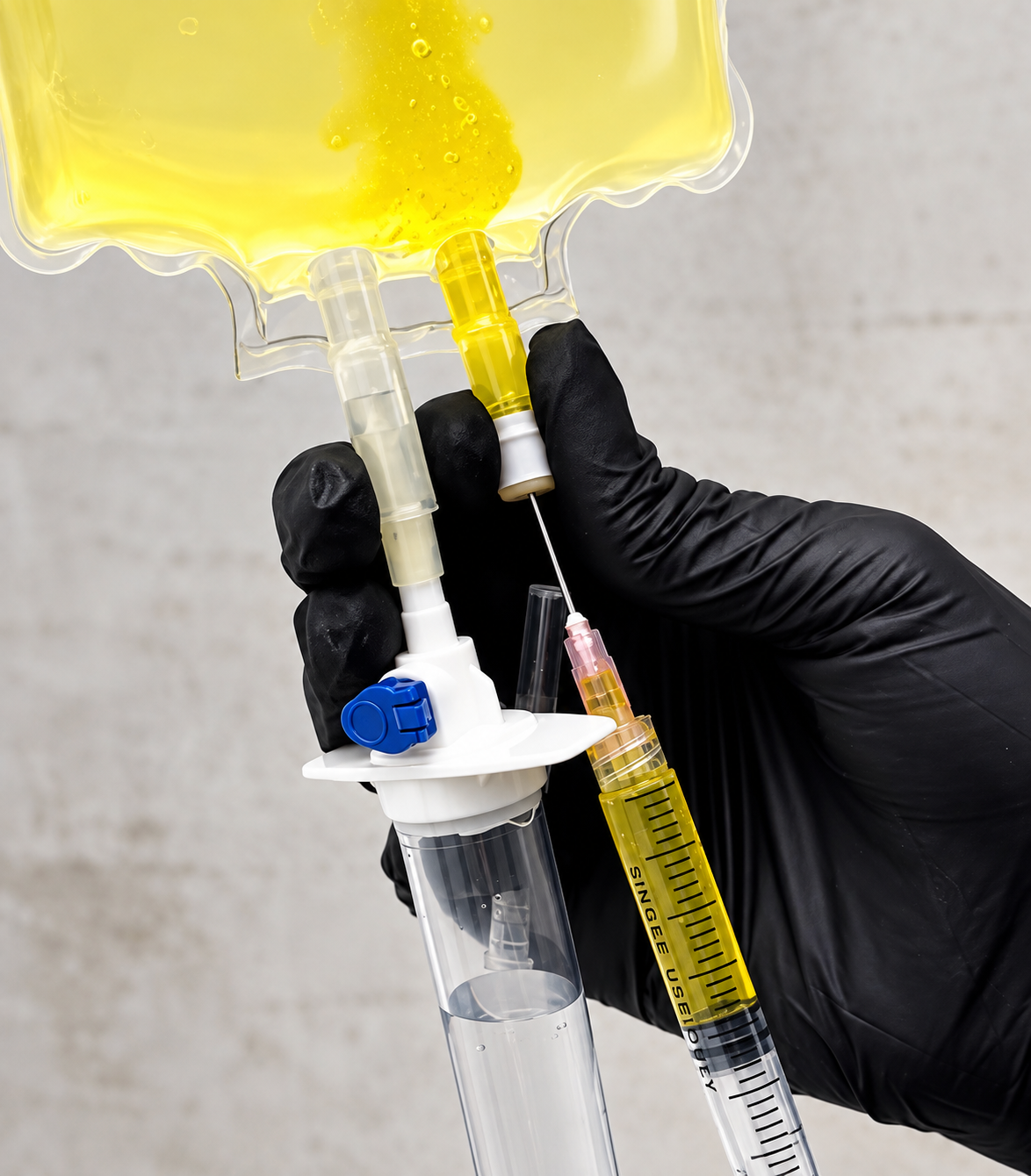 A hand wearing a black glove holding a syringe and a vial of yellow liquid, with a large container of yellow liquid in the background.