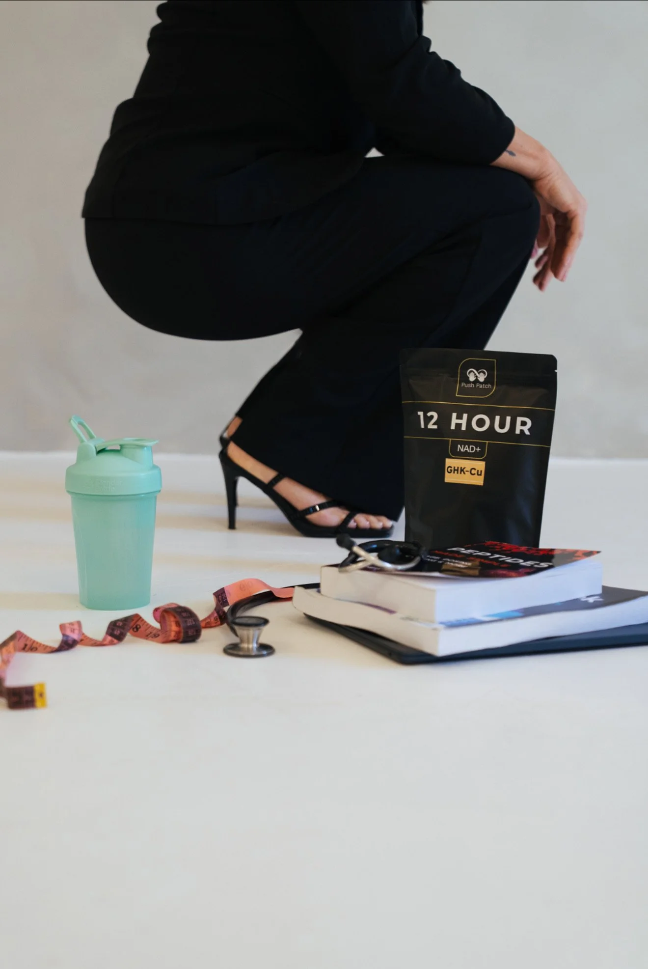 A person dressed in black squatting on the floor, with various items including a mint green shaker bottle, a measuring tape, books, and office supplies on a white surface in front of them.