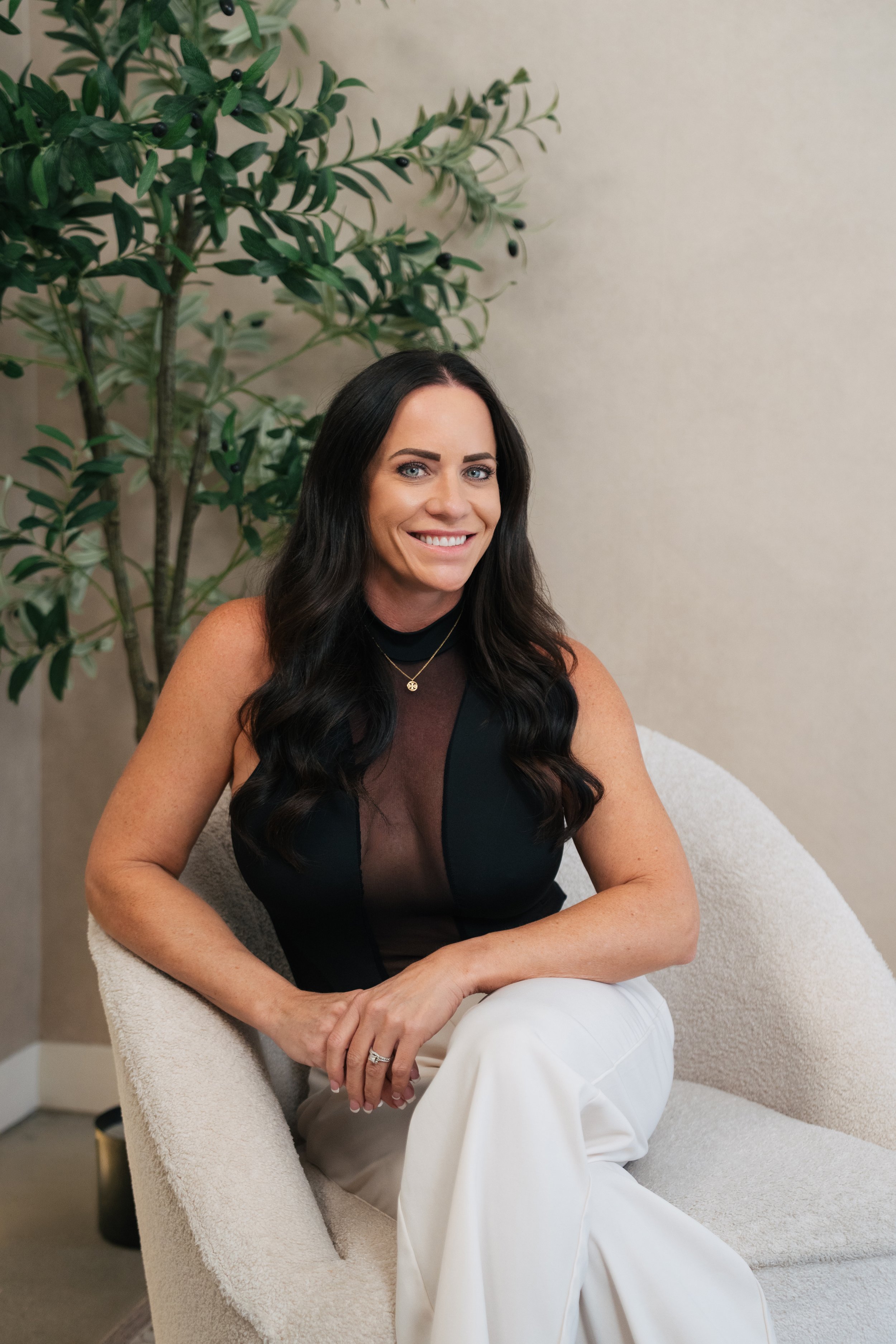 A woman with long dark hair, wearing a black sleeveless top and white pants, sitting in a light-colored chair next to a green plant with small black berries, smiling at the camera.