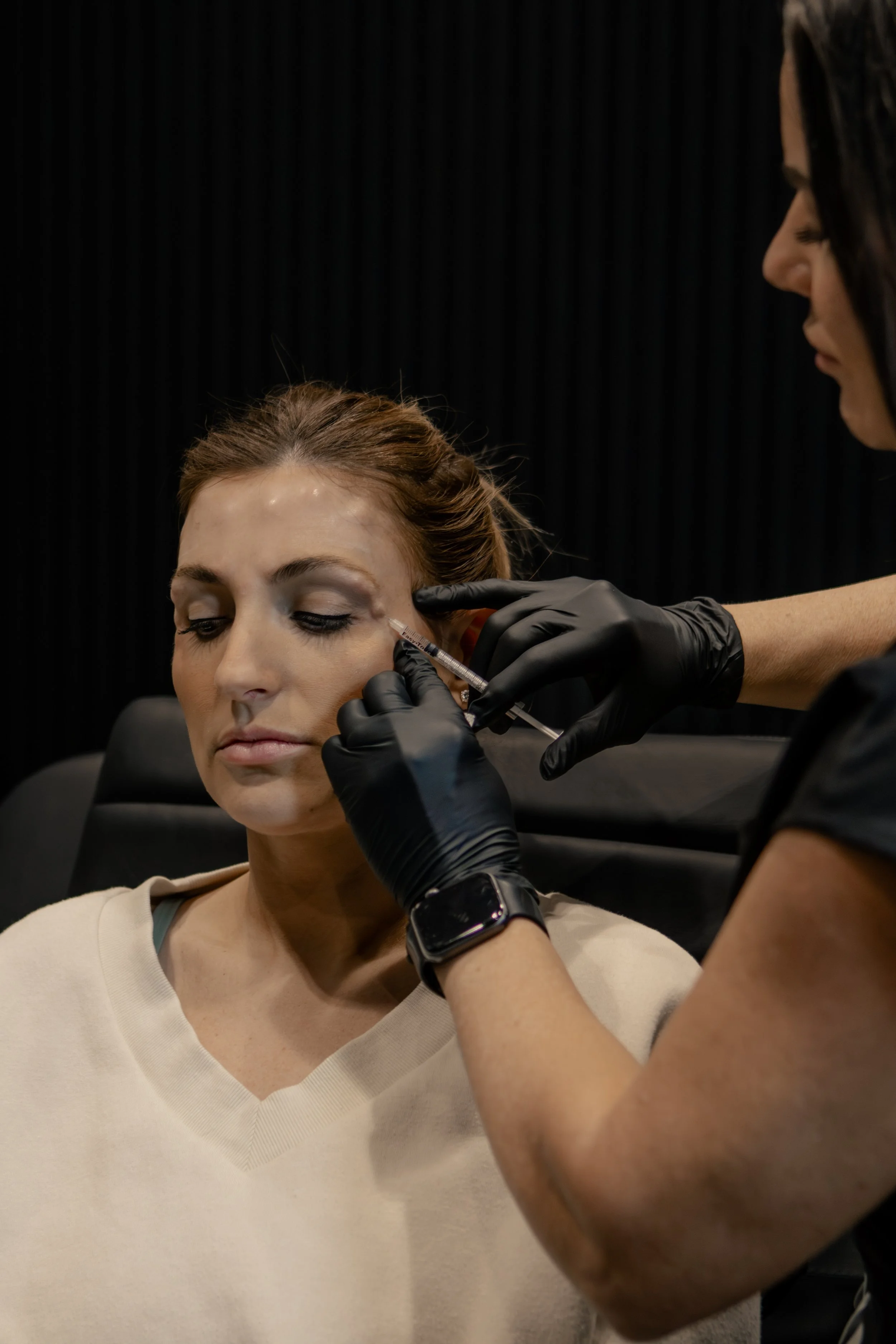 A woman with closed eyes receiving a cosmetic injection under her left eye from a professional wearing black gloves.