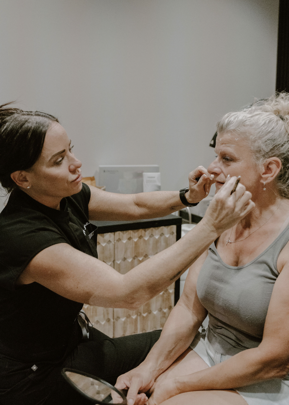 A makeup artist applying makeup to an elderly woman with gray hair in a salon or spa setting.
