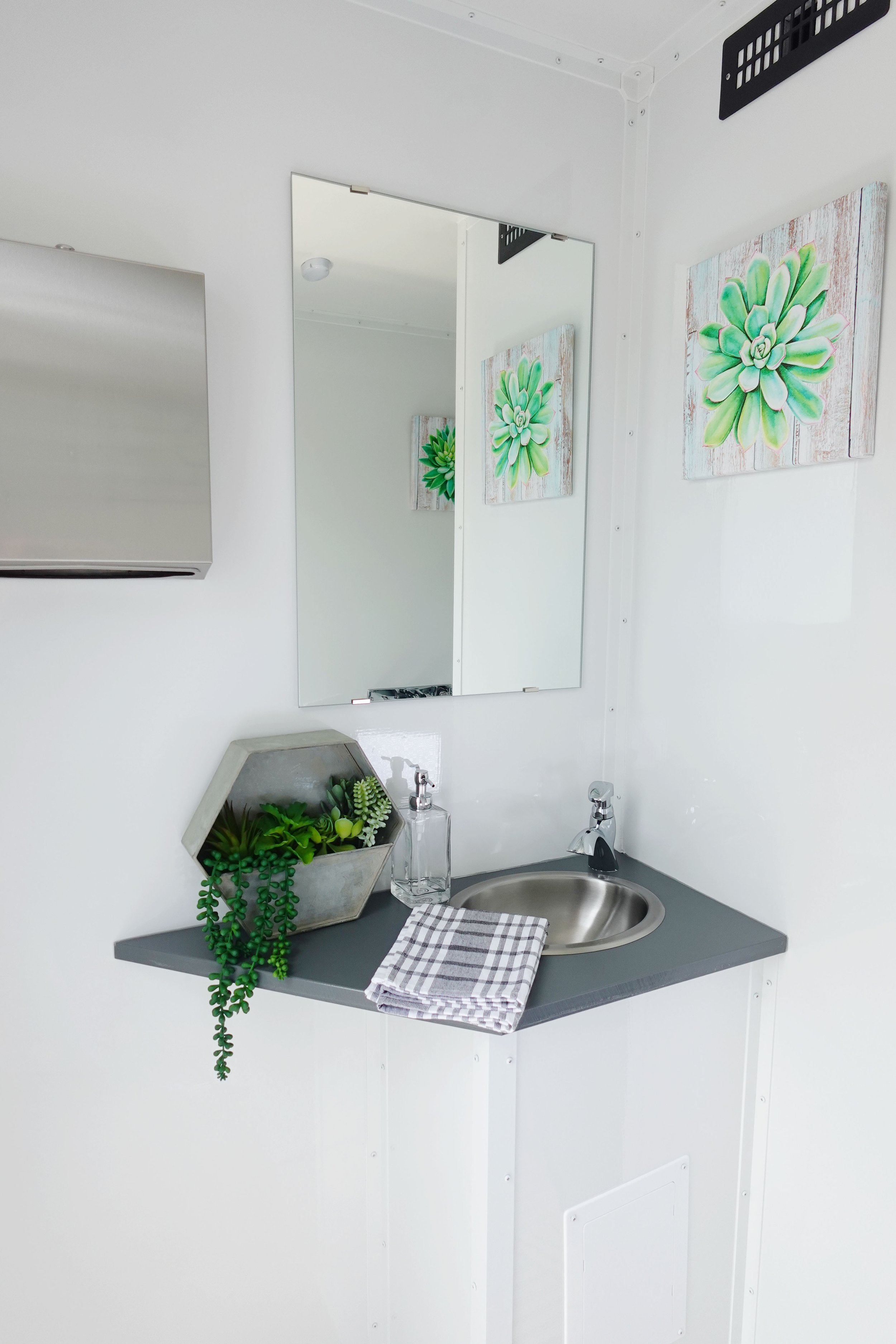 A small workspace with a gray countertop, a round stainless steel sink, a black faucet, and a striped cloth towel. There is a hexagonal concrete pot with green plants and a glass soap dispenser. A foldable napkin is on the counter. A rectangular mirror is above the sink, with two paintings of green succulent plants hanging on the white wall behind. A black vent cover is on the ceiling, and a white fire alarm is visible in the mirror.