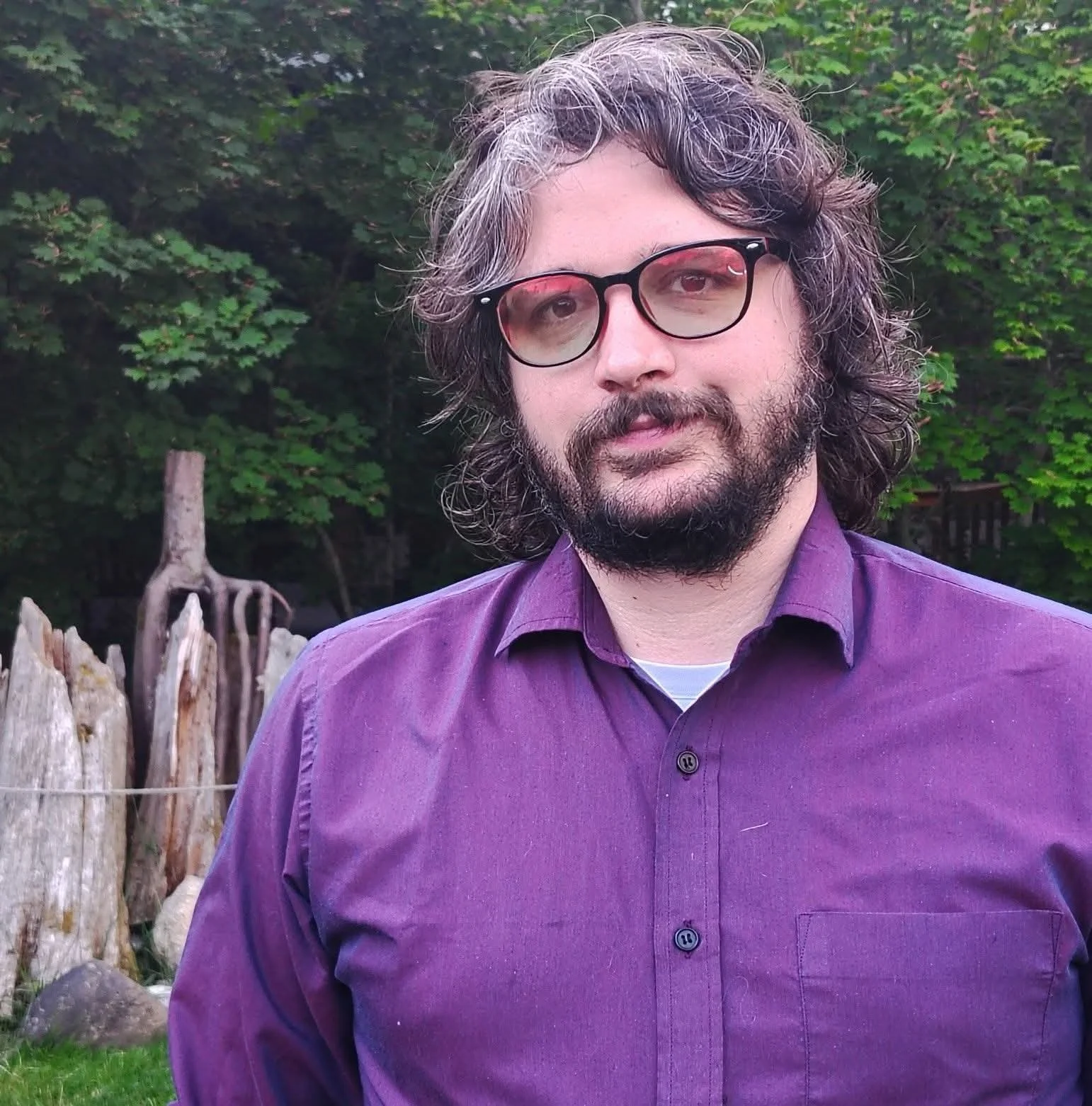 A man with long, curly hair and a beard wearing glasses and a purple shirt, standing outdoors with green foliage in the background.