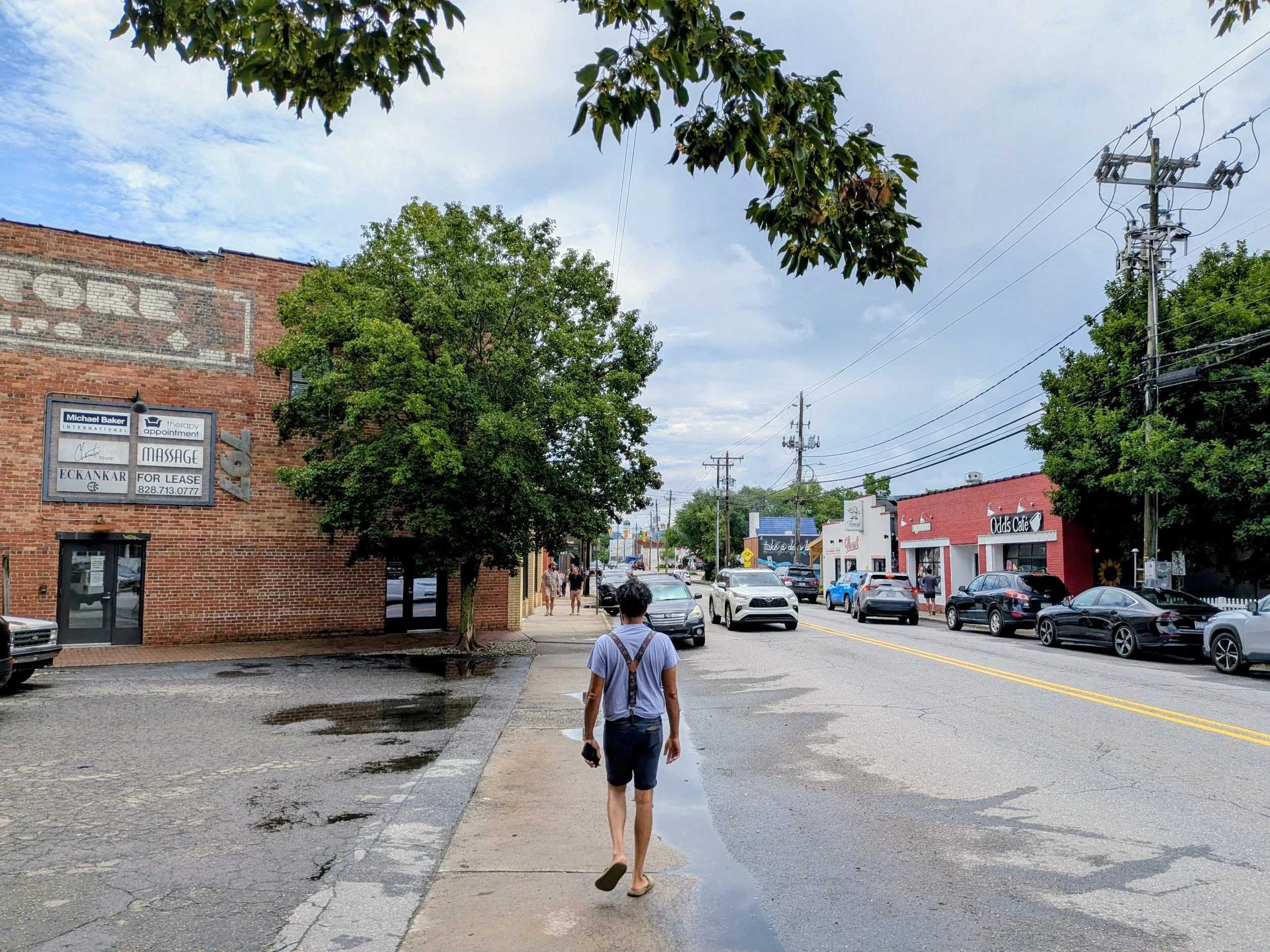 A young person walking on a sidewalk in Haywood Rd in West Asheville, a small-town area, with cars parked along the street and buildings, trees, and utility poles in the background. The sky is partly cloudy, and there are puddles on the pavement.