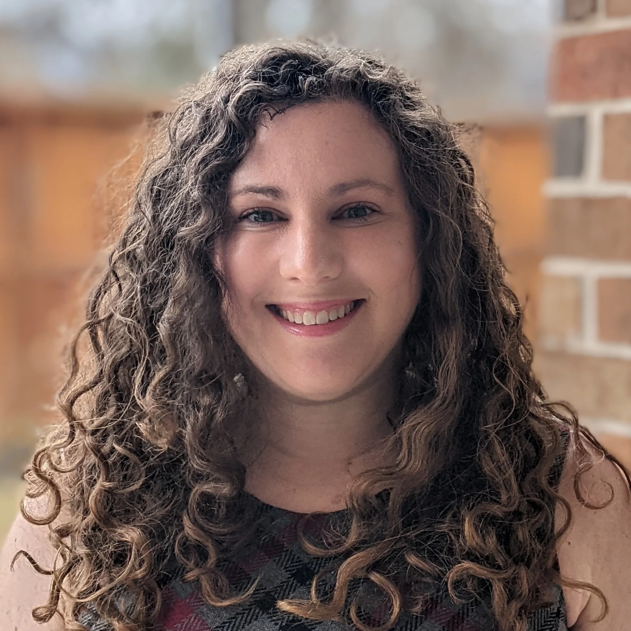 Close-up of a woman with curly brown hair, smiling, with bricks and a wooden surface in the background.