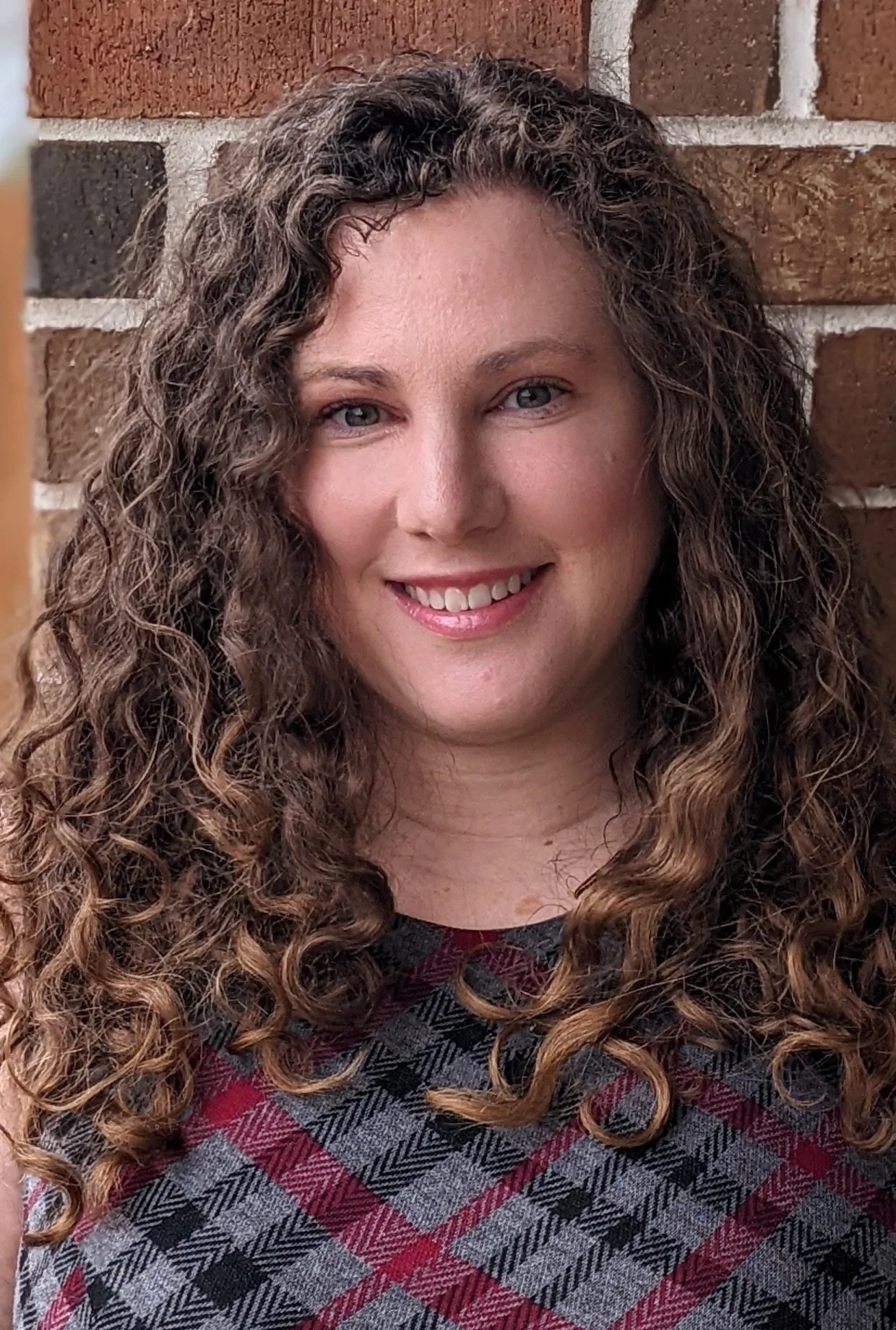 A woman with long, curly brown hair and blue eyes smiling in front of a brick wall.