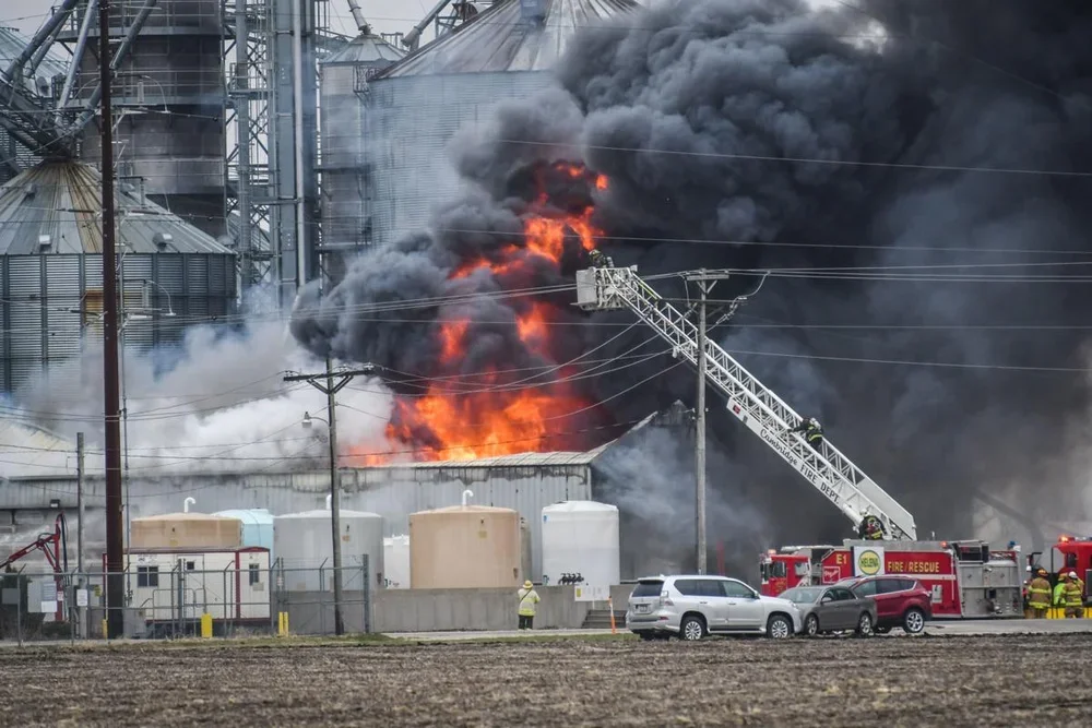 Industrial building on fire with large black smoke and flames, firefighters using a ladder truck to fight the fire, several emergency vehicles and personnel present.