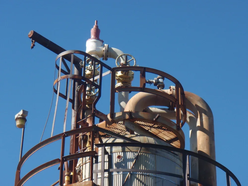 Close-up of an industrial distillation tower with pipes, valves, and walkways against a clear blue sky.