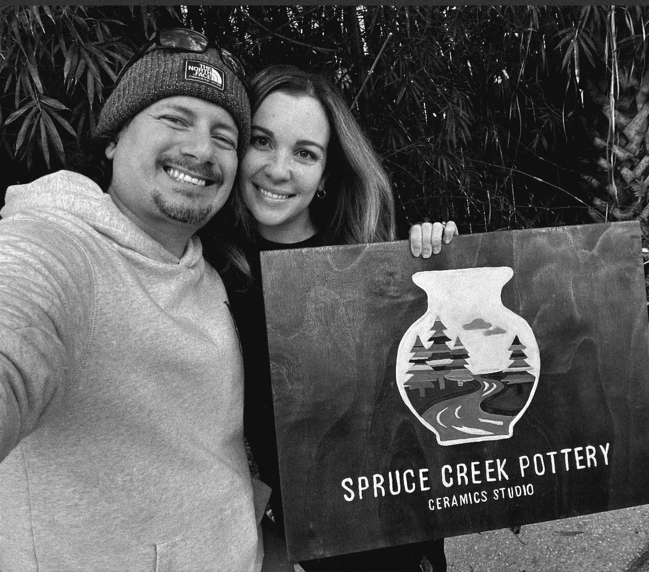 A man and a woman smiling and taking a selfie. The woman holds a sign that says 'Spruce Creek Pottery, Ceramics Studio' with a graphic of a vase, trees, a river, and a cloud.