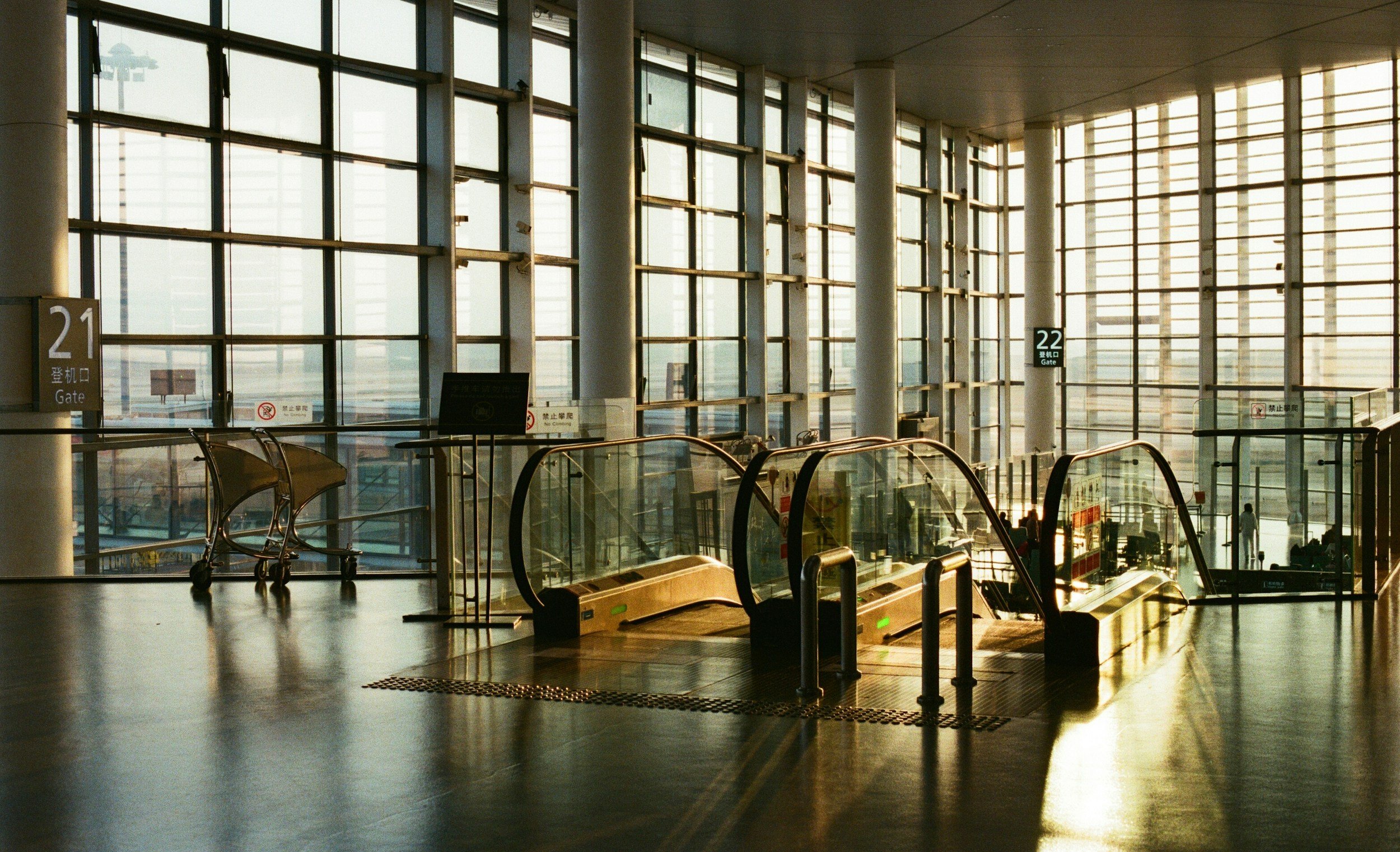 Inside an airport terminal showing large glass windows, escalators, and empty seats near gate 21 and 22, with bright sunlight streaming in.