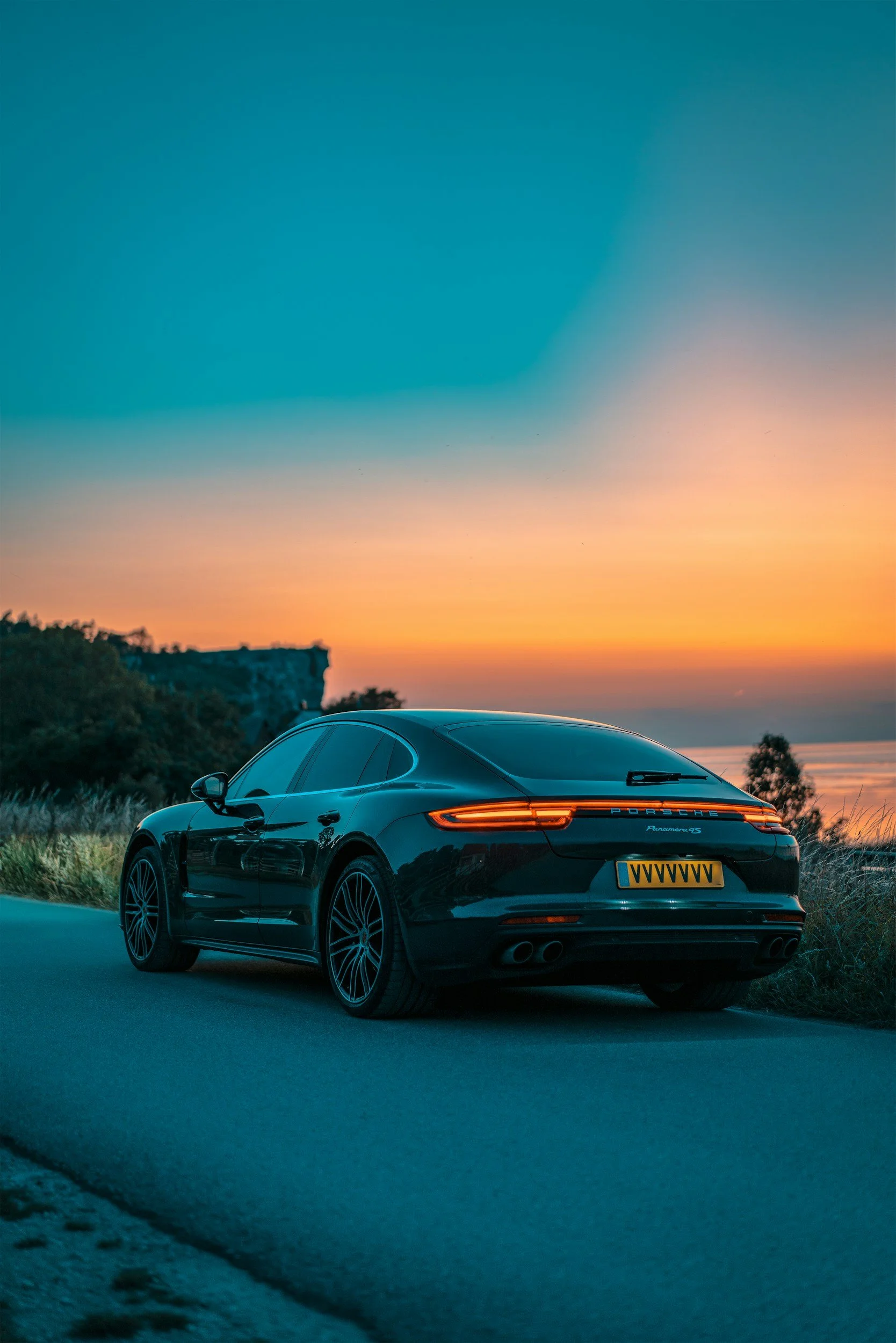 A black Porsche Panamera 4S with yellow license plate parked on a road during sunset, with a sunset sky and distant trees in the background.