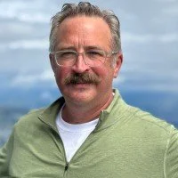 A middle-aged man with glasses and a mustache, wearing a green jacket over a white shirt, standing outdoors with a cloudy sky in the background.
