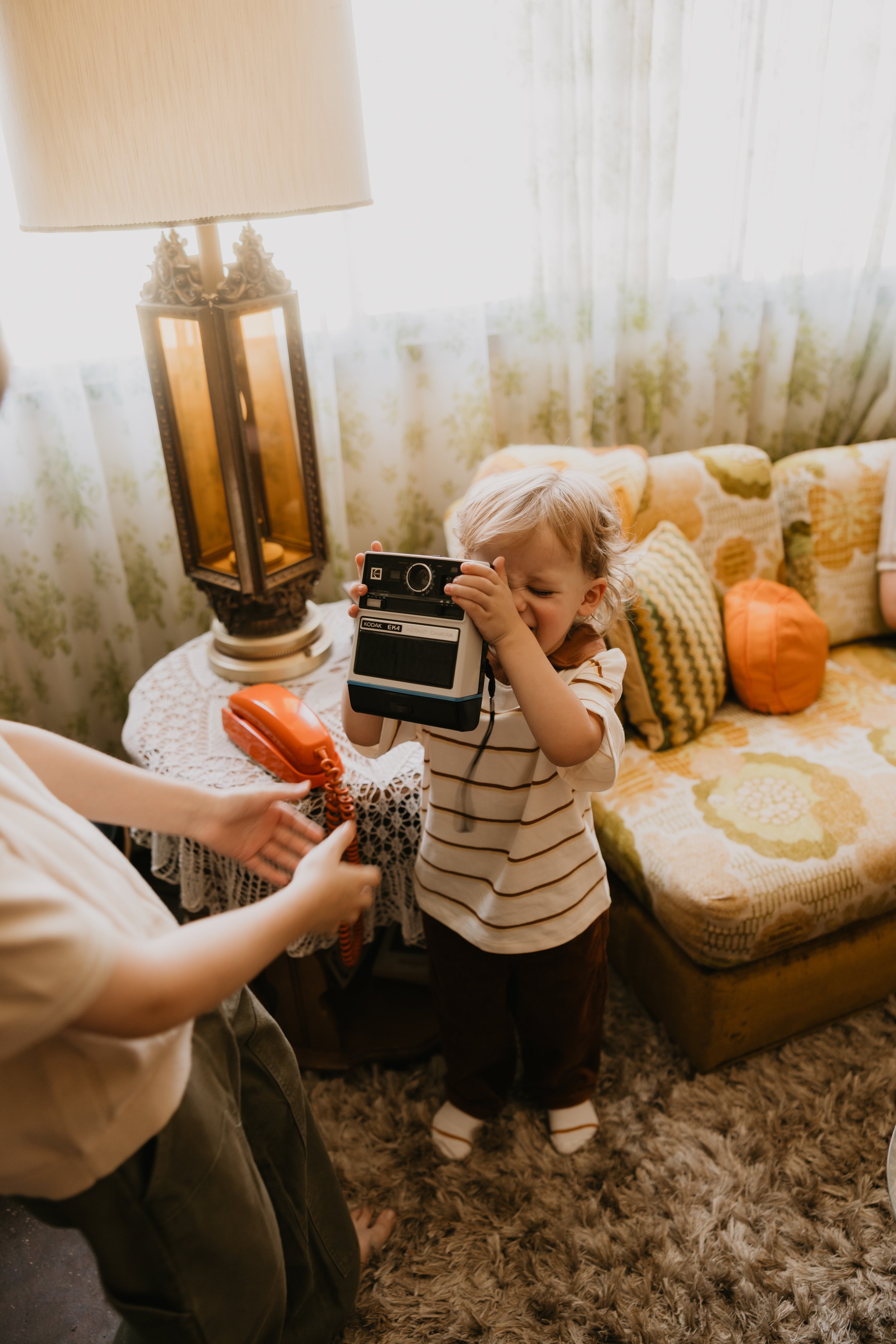 A young child with curly blond hair is looking at an instant camera and laughing, with people around him holding a vintage orange telephone. The scene is set in a living room with a floral patterned couch, several throw pillows, and a lamp next to a window.