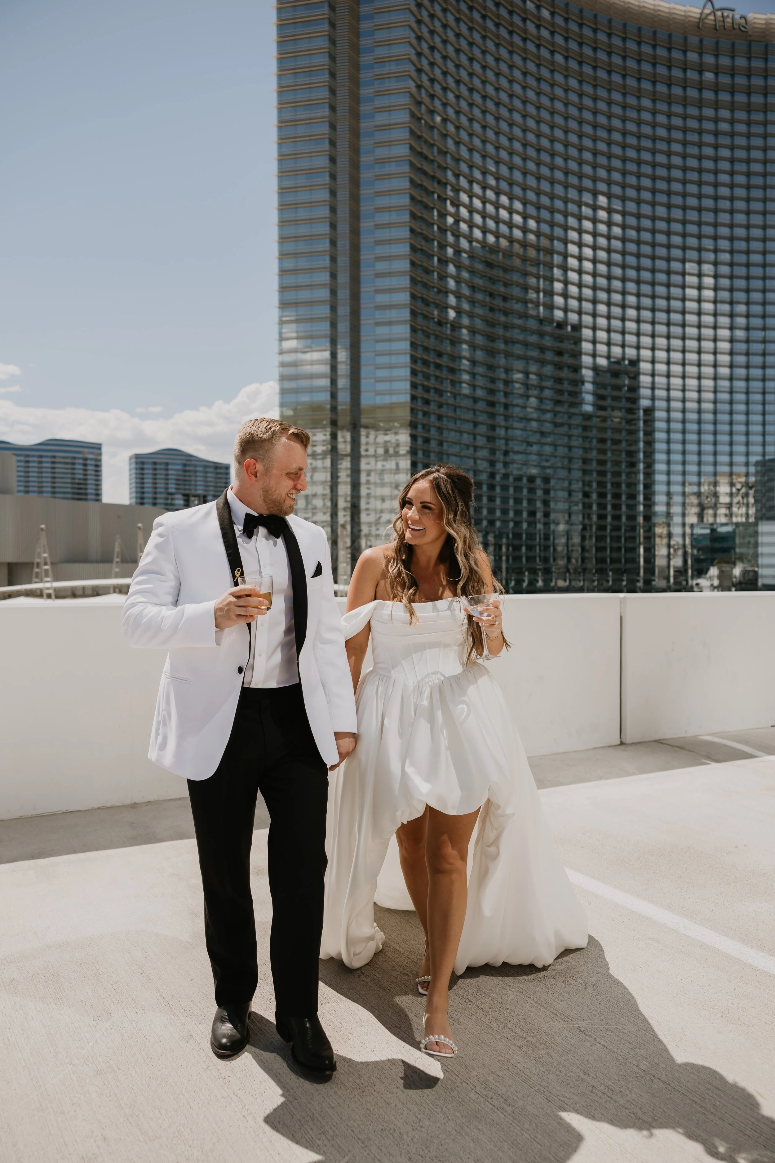 A man and woman dressed in wedding attire walking and laughing together on a rooftop parking lot with a city skyline in the background. The man is wearing a white tuxedo jacket with black lapels, a black bow tie, and black trousers. The woman is in a white strapless wedding dress with a full skirt, holding a glass, and wearing high-heeled sandals.