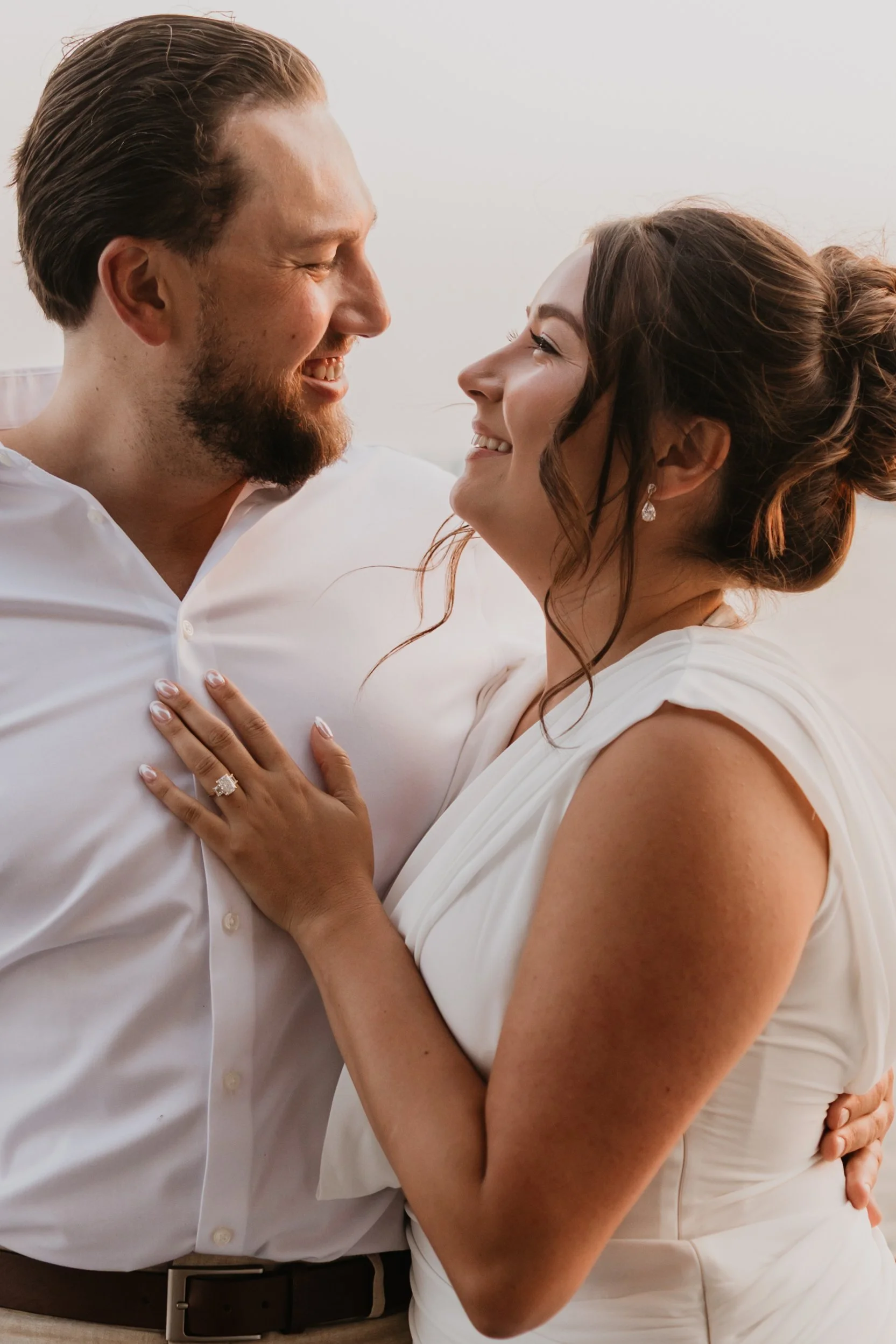 A couple smiling and looking into each other's eyes, with the woman wearing wedding rings and earrings.