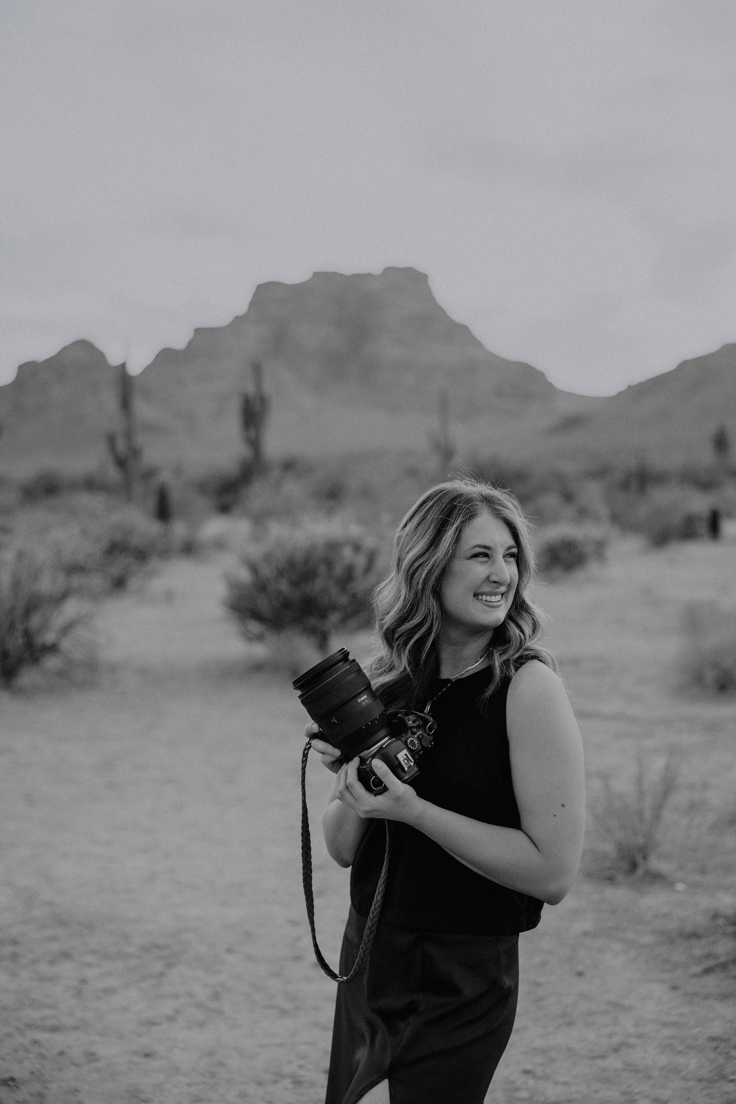 Black and white photo of a woman smiling outdoors in a desert landscape, holding a camera. There are cacti and mountains in the background.