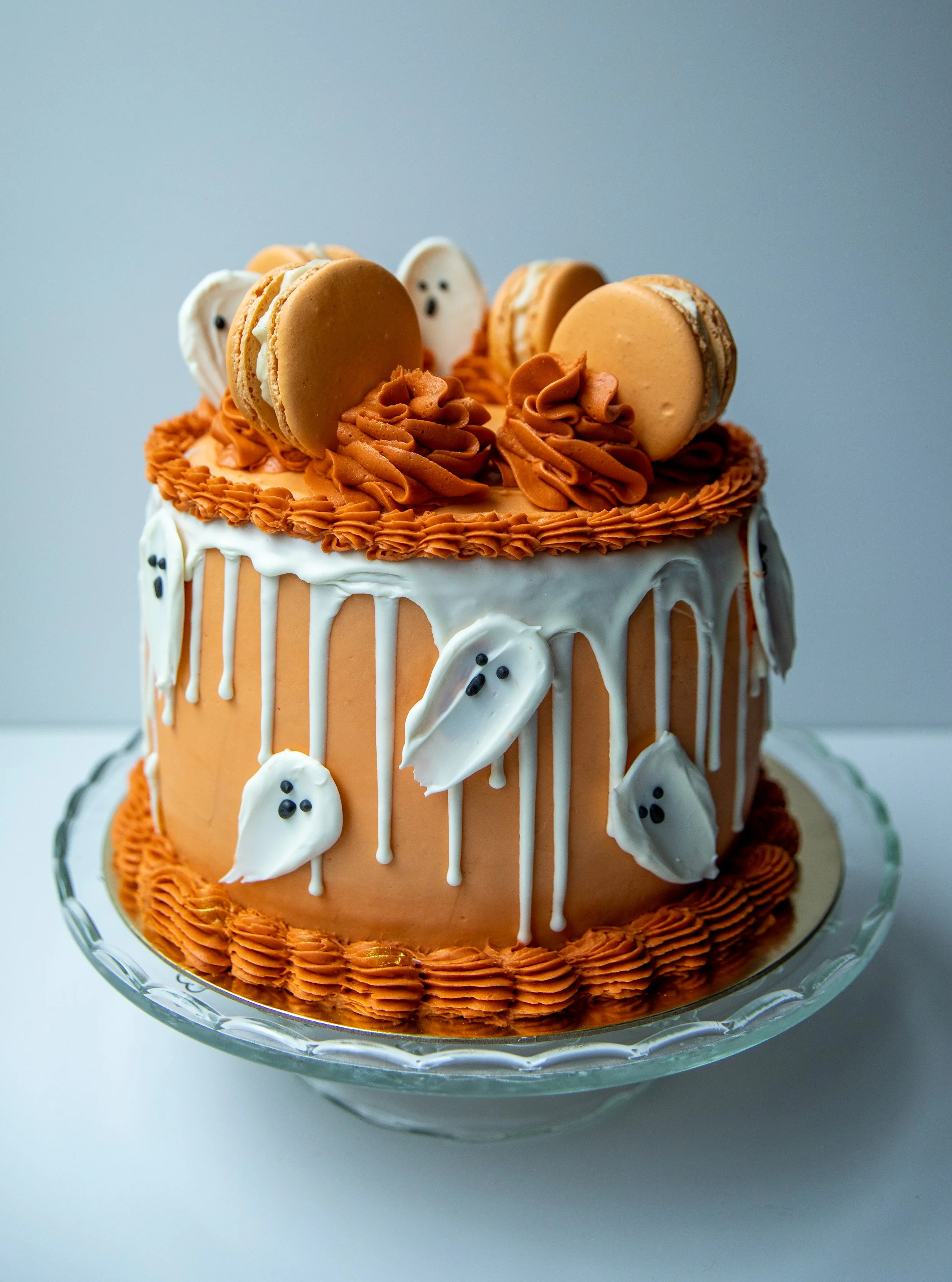 A Halloween-themed cake decorated with orange, white, and black icing, featuring piped orange frosting on top and ghost-shaped decorations with black eyes and mouths, topped with macarons in orange and beige colors.