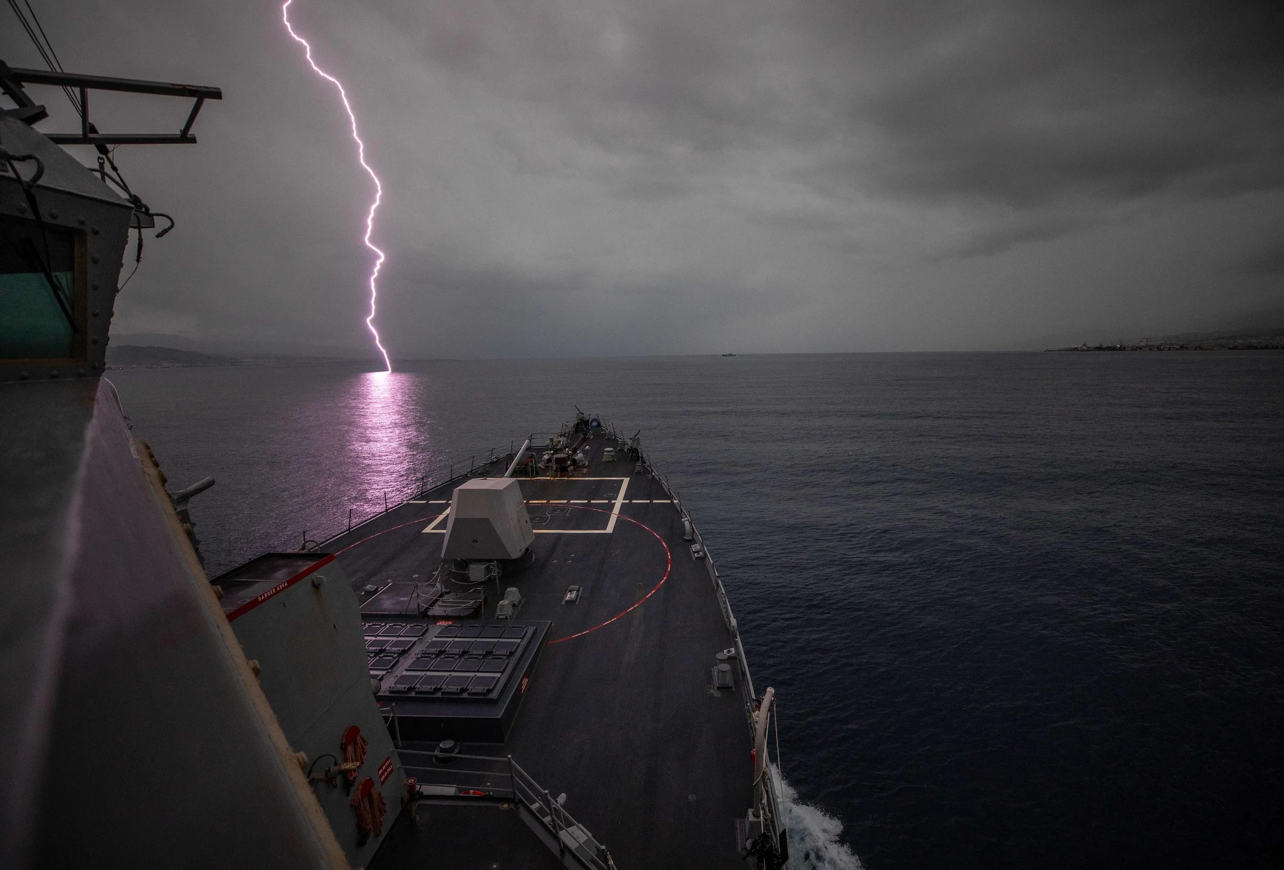 A naval ship at sea during a thunderstorm with visible lightning bolt in the cloudy sky.