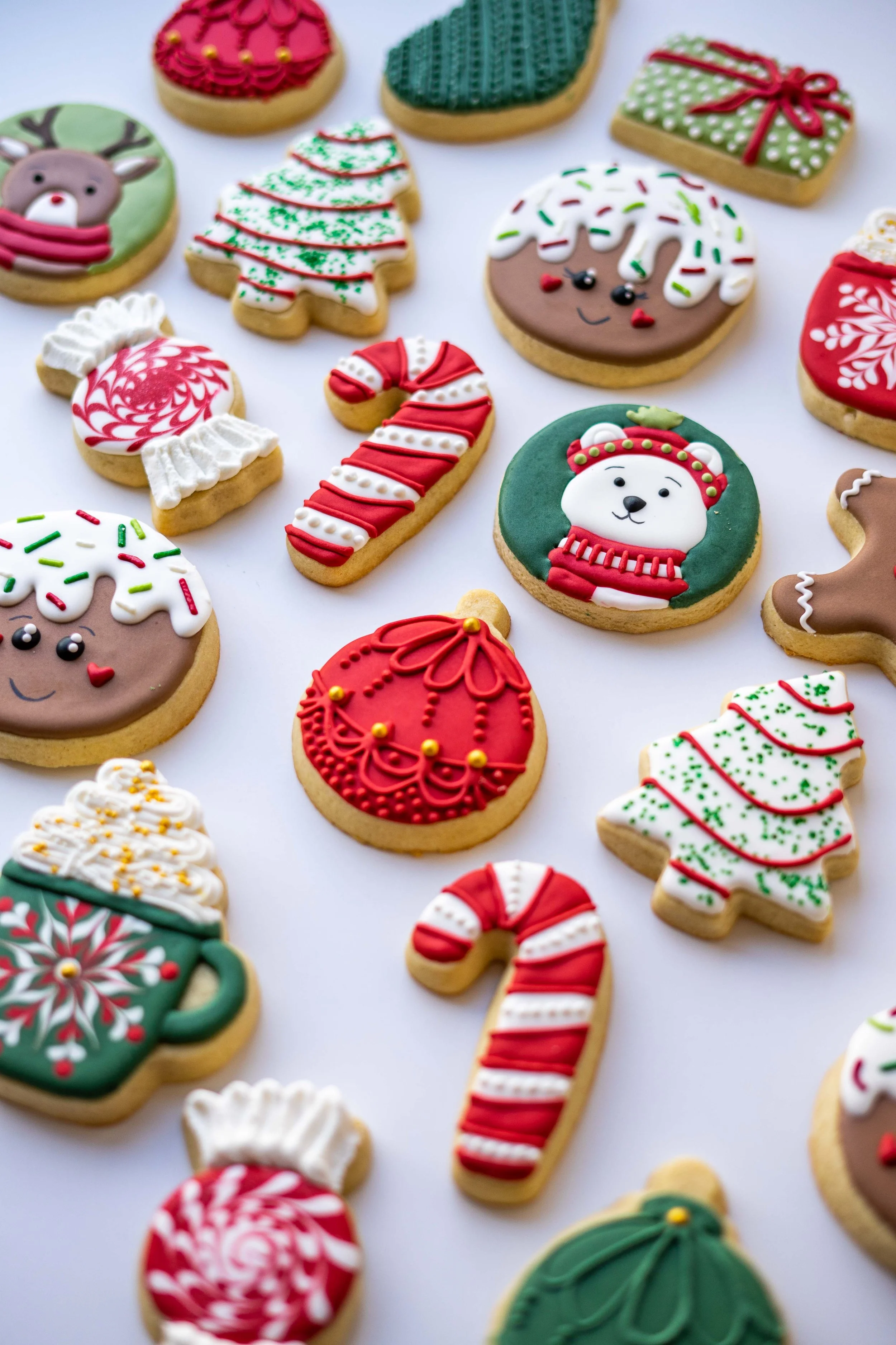 Assorted decorated Christmas cookies, including a reindeer, Christmas tree, snow-covered house, candy cane, snowflake, snowman, and gift box, all with festive colors and icing.