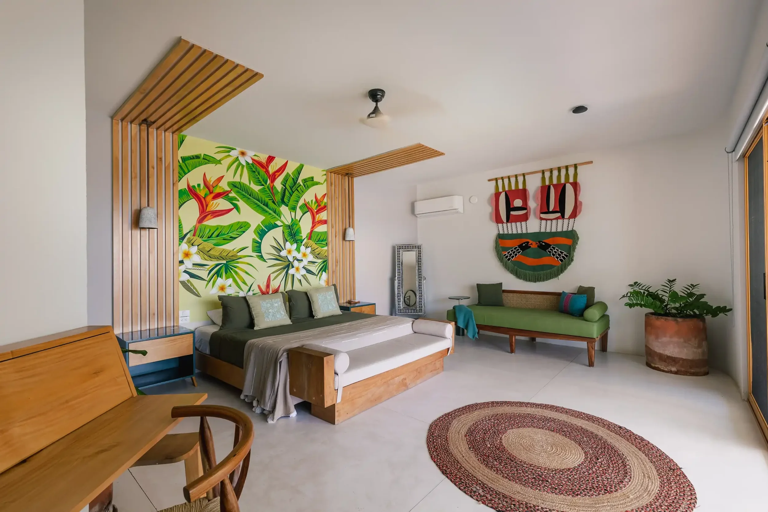 Living room with a tropical-themed wall mural, wooden accents, green sofa, rug, framed mirror, and decorative textiles, illuminated by natural light from large windows.