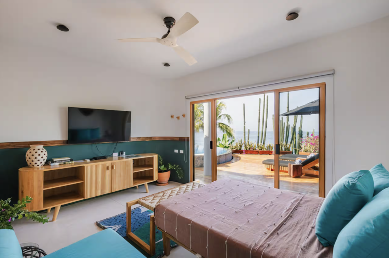 Bedroom with a sliding glass door leading to a balcony with outdoor furniture and a view of cacti and palm trees.