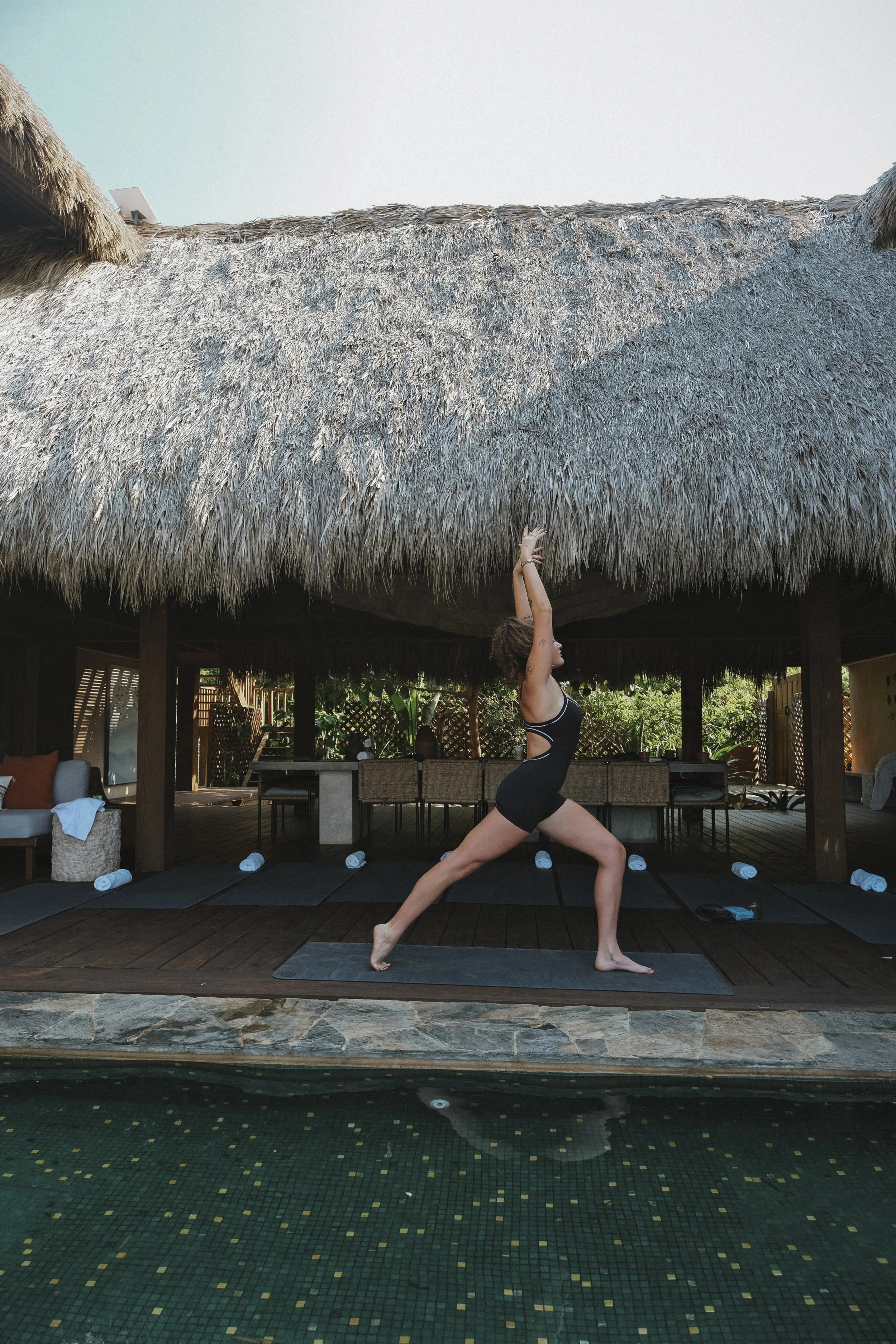 A woman doing a yoga pose on a yoga mat under a thatched roof, next to a pool with a tiled floor, in a tropical outdoor setting.