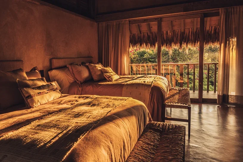 Two beds with pillows in a rustic bedroom with an open balcony showing lush greenery outside at sunset.