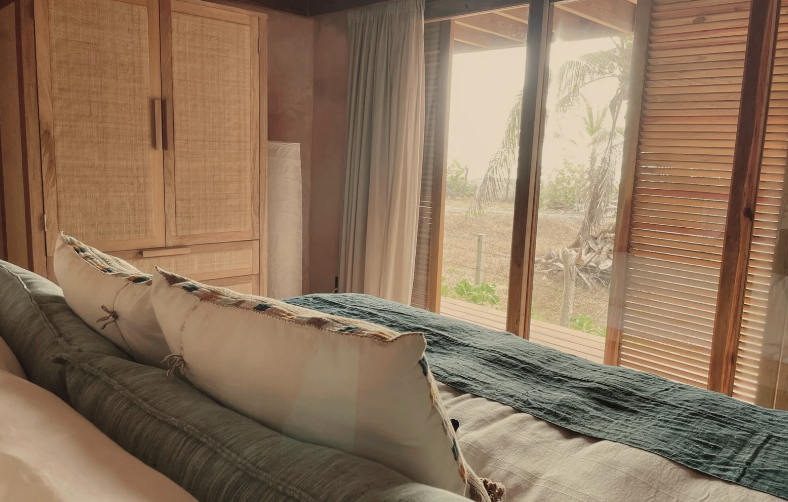 Bedroom with large sliding glass doors showing outdoor trees, wooden window shutters, a bed with pillows and a green throw blanket, and wooden cabinets.