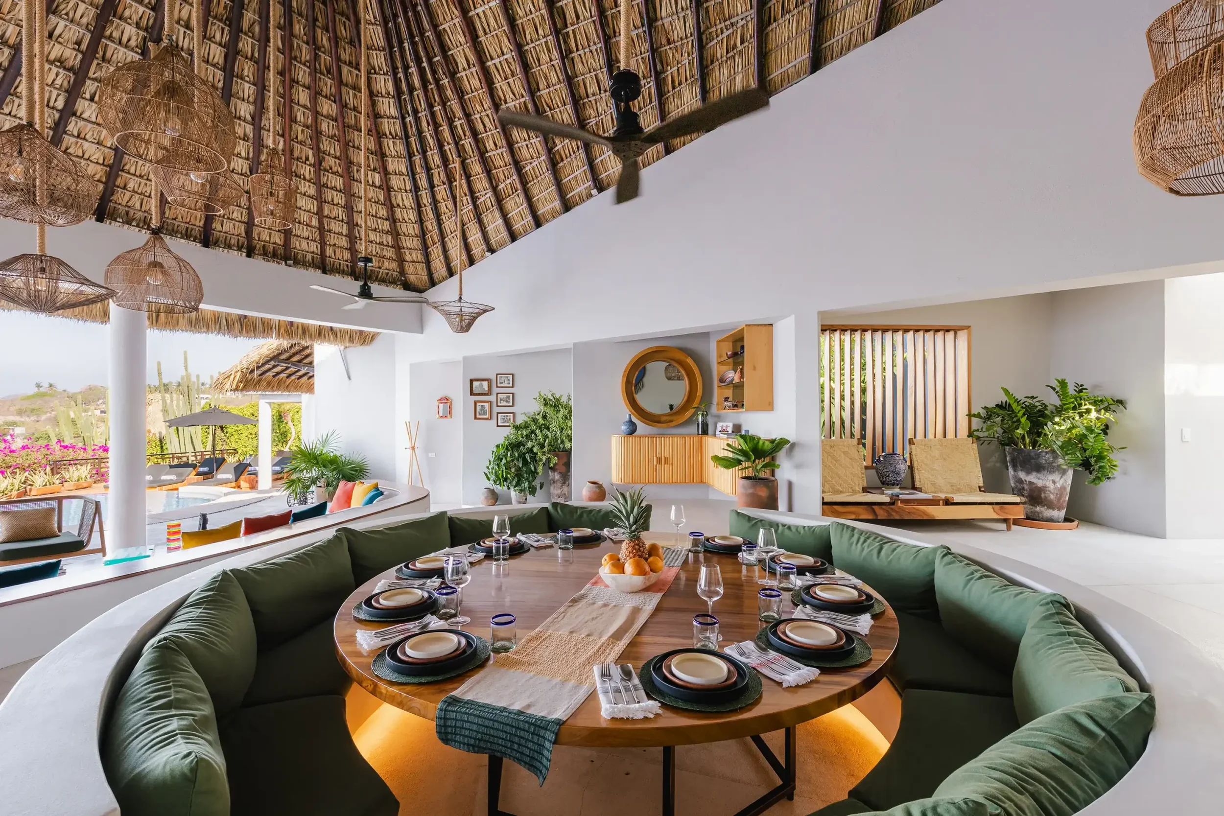 A spacious indoor dining area with a round wooden table set with plates, glasses, and a fruit centerpiece. There is a large, curved sofa with green cushions around the table. The room features a high, thatched ceiling with ceiling fans and hanging woven light fixtures. Walls are white with wooden accents, and large windows with views of an outdoor patio and landscape.