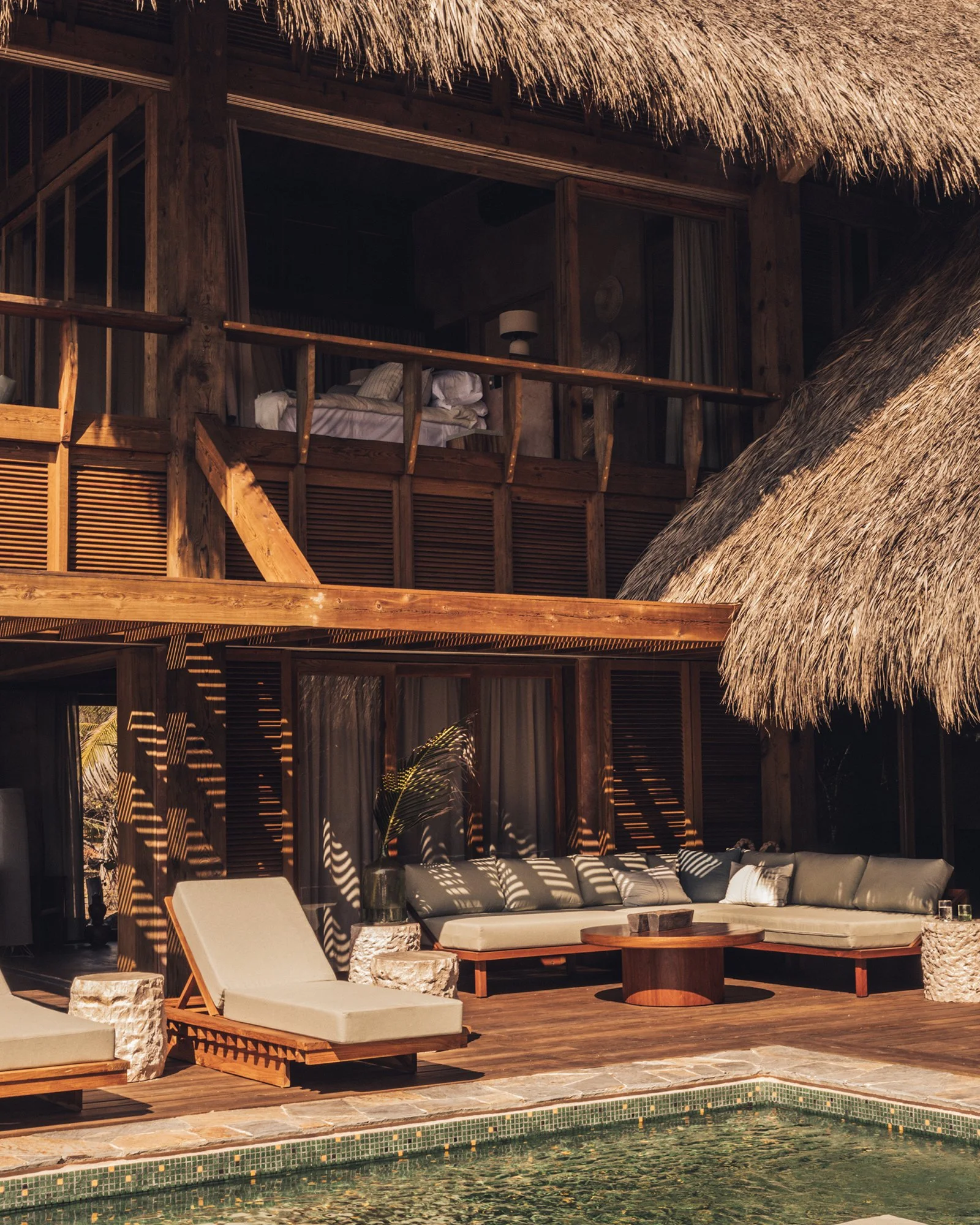 Luxury resort pool area with wooden sun loungers, a white sectional sofa, and a thatched roof structure, surrounded by lush greenery.