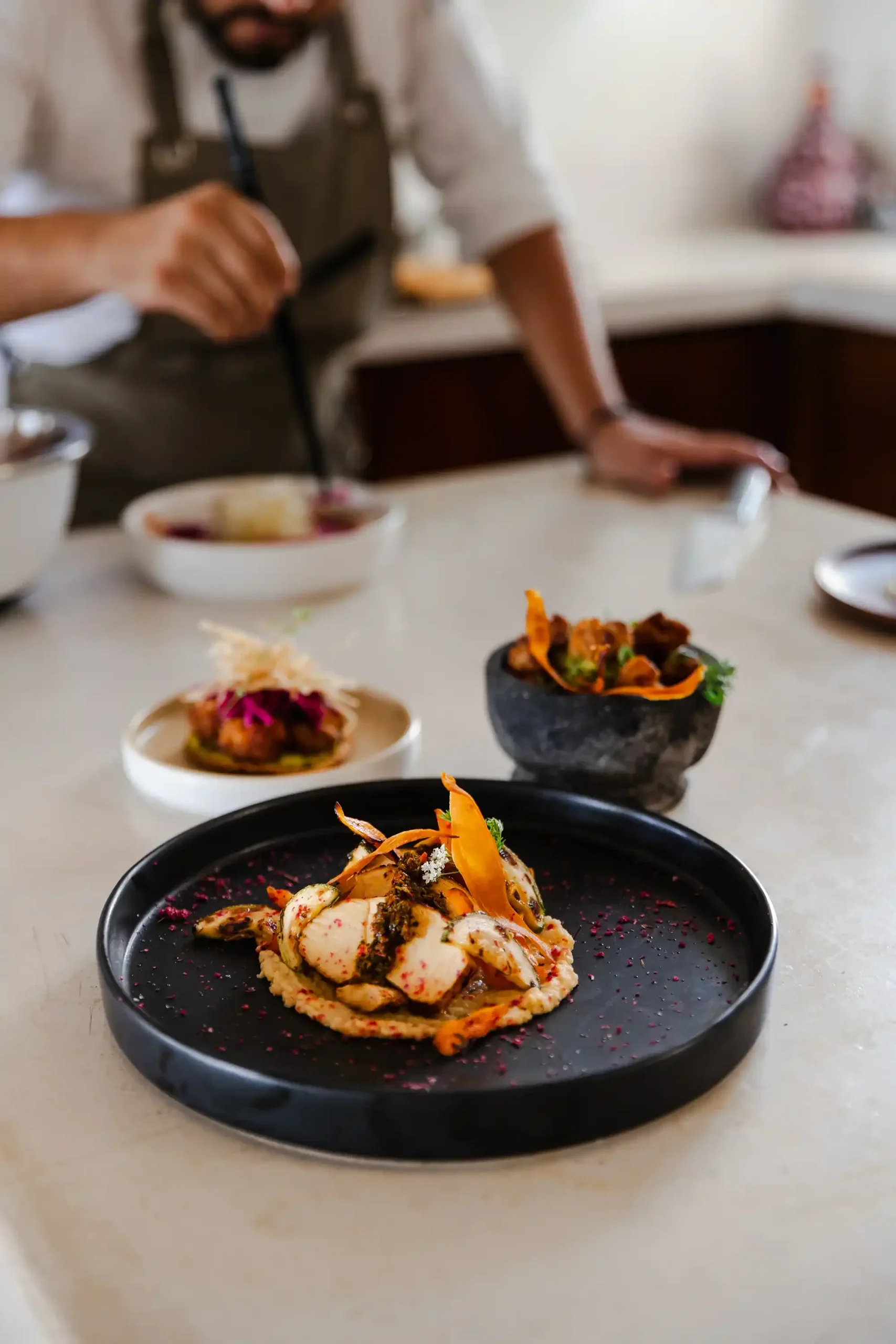 Close-up of a black plate with a gourmet dish topped with herbs and vegetables on a white table, with blurred background showing other dishes and a person preparing food.