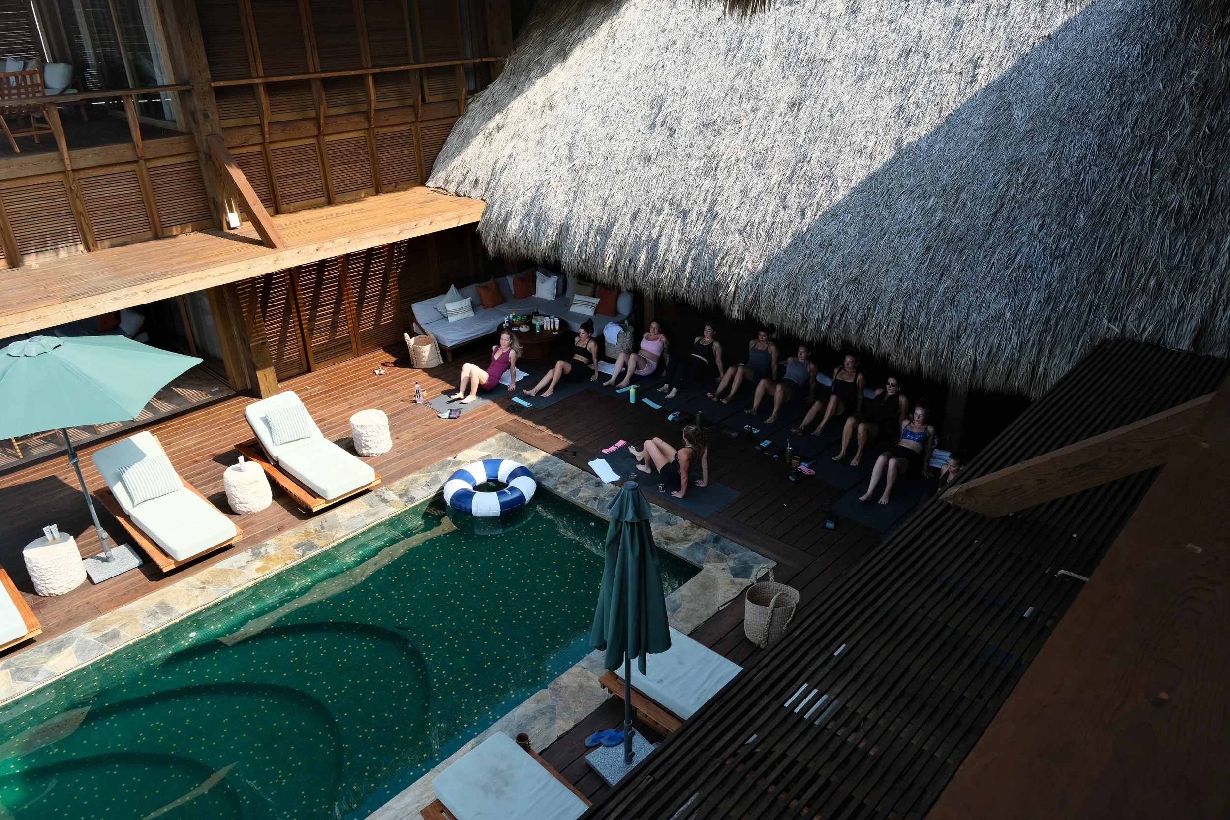 People practicing yoga on a deck beside a swimming pool in an outdoor area with thatched roof and wooden structures.