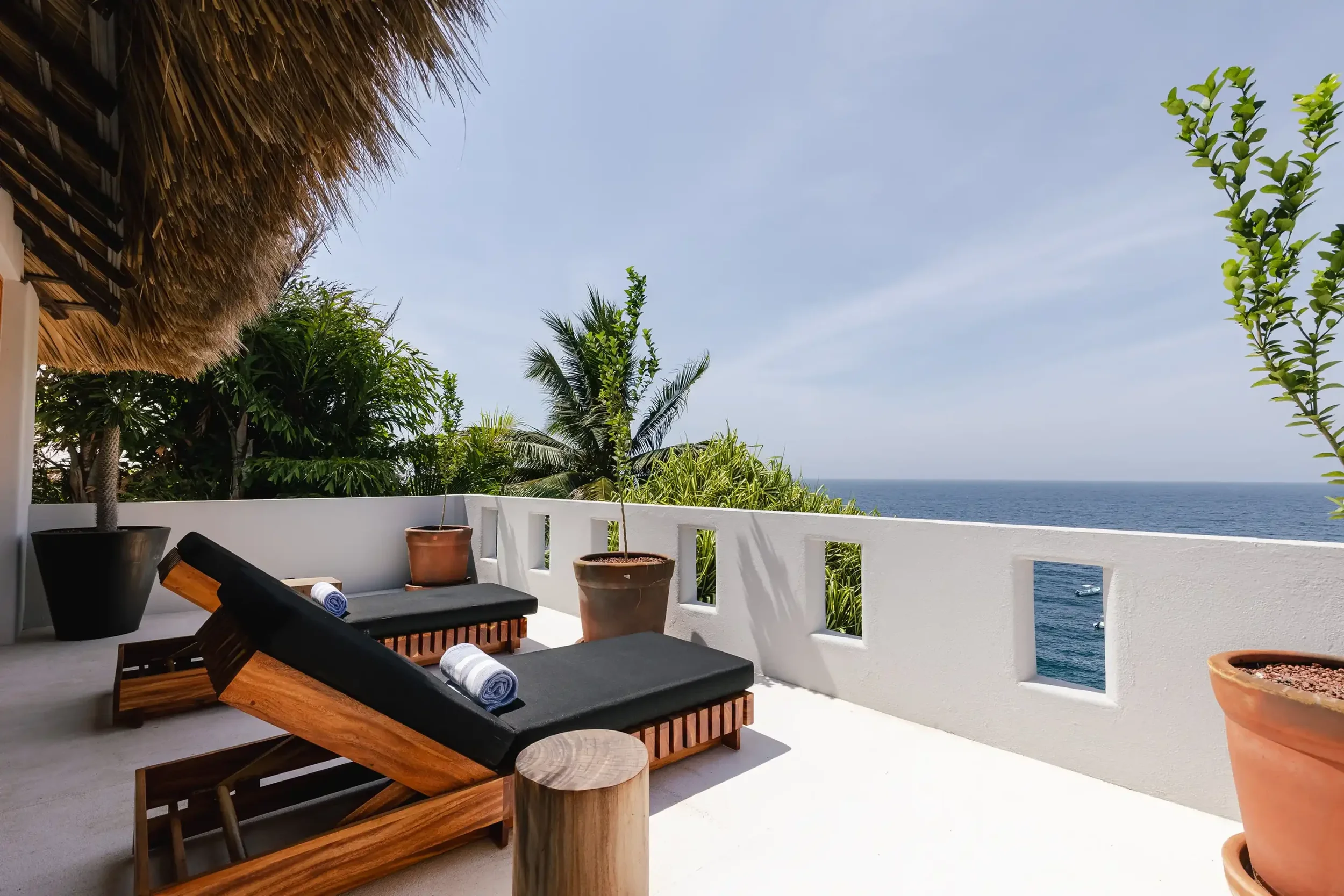 A balcony with two black lounge chairs, rolled towels, a small round wooden table, and potted plants, overlooking the ocean with clear skies and lush green trees nearby.