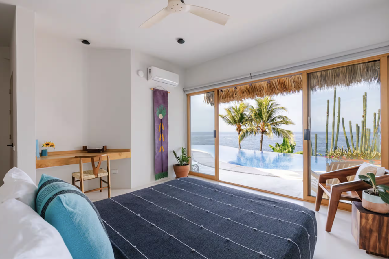 Bedroom with large sliding glass doors overlooking a pool and ocean, decorated with tropical plants, a ceiling fan, and minimalist furniture.
