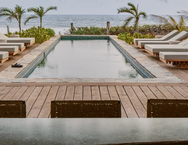 Poolside view with lounge chairs, palm trees, and ocean in the background.