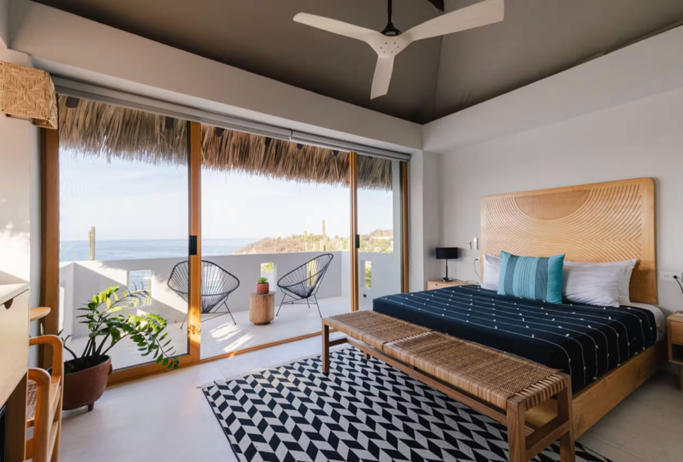 A beachside hotel room with a large sliding glass door leading to a balcony with two black chairs and a small table, overlooking the ocean. The room features a wooden bed with a geometric headboard, a black and white patterned rug, and a plant in a pot near the door.