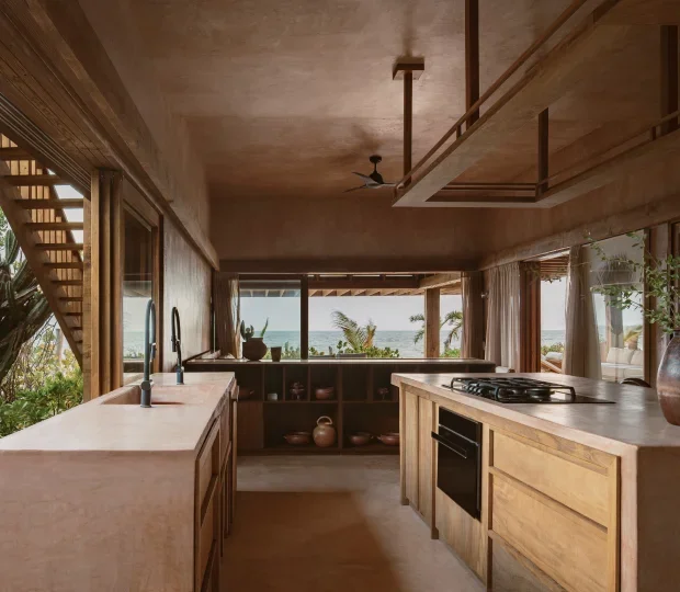 Modern kitchen with wooden cabinetry, facing large windows with a view of trees and the ocean, featuring a marble island, built-in oven, and a gas stove, decorated with plants and pottery.