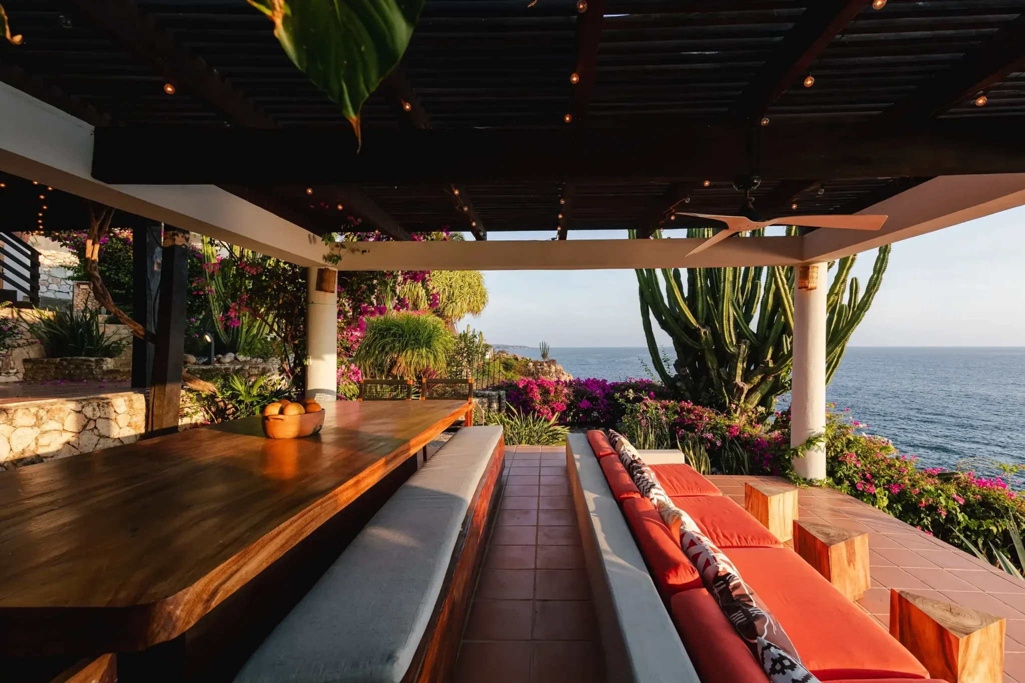 Outdoor patio with wooden dining table and cushioned seating, surrounded by pink and purple flowering plants, overlooking the ocean under a dark pergola with string lights and a ceiling fan.