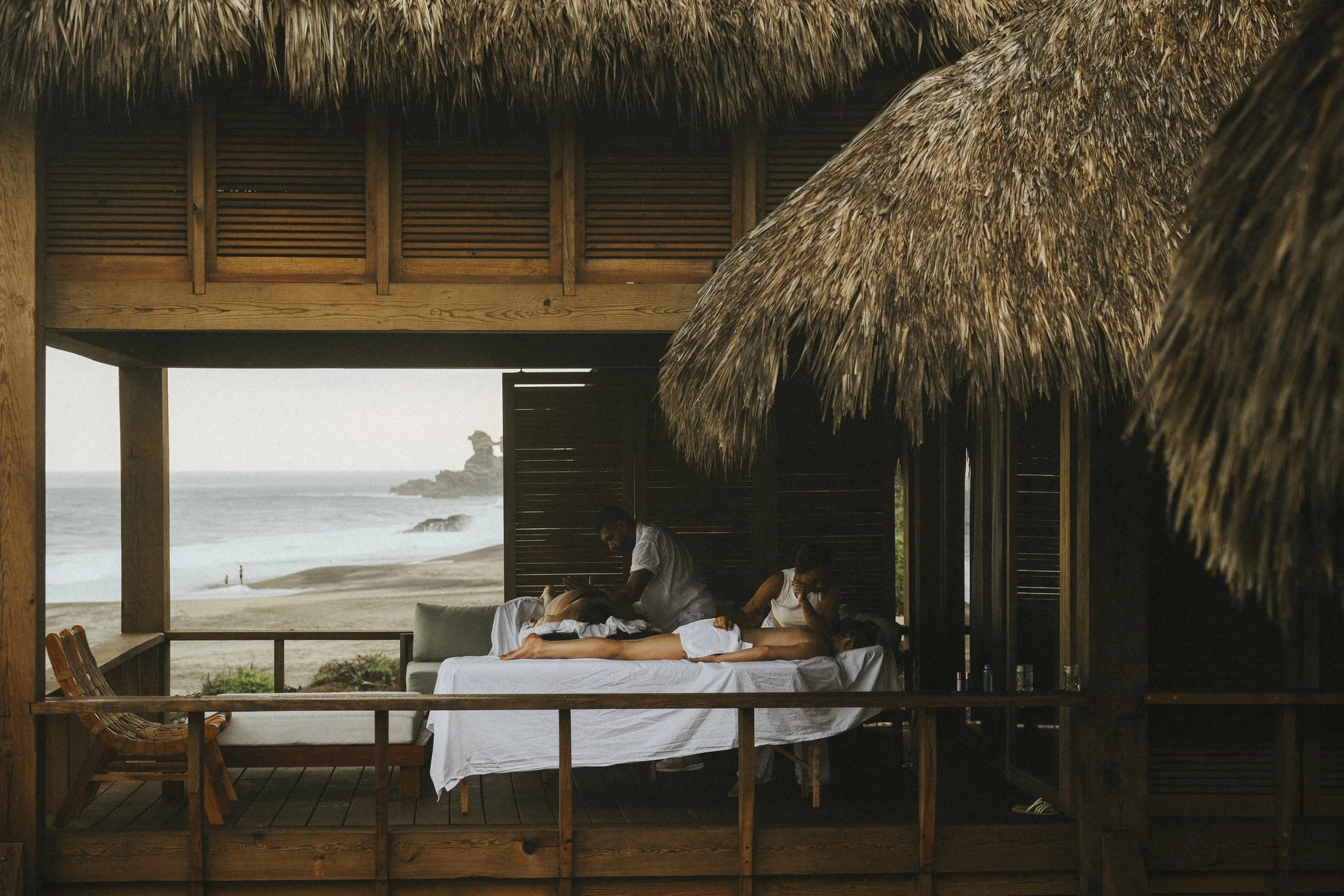 A couple getting a massage on a bed under a thatched roof with a view of the beach and ocean in the background.