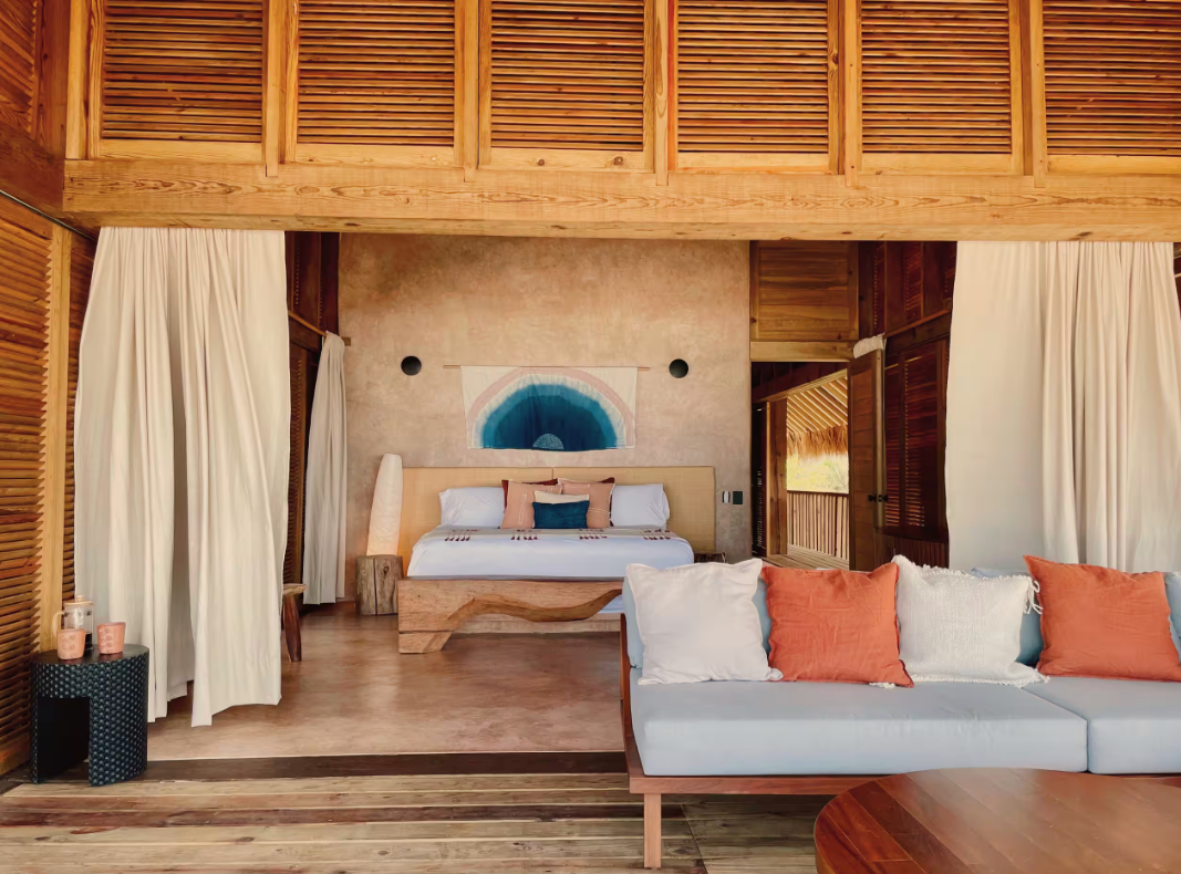 A spacious bedroom with a king-sized bed featuring white bedding and pillows, a rustic wooden bench at the foot of the bed, and an abstract blue wall hanging behind it. There are white curtains on each side, a seating area with white and orange pillows, a small black side table with candles, and wooden accents on the walls and ceiling.