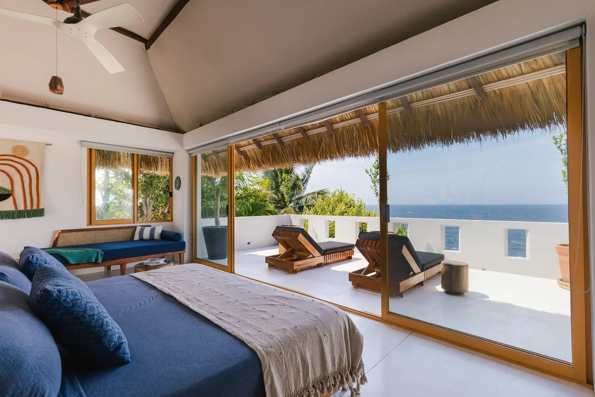 Living room with sliding glass doors leading to a balcony overlooking the ocean, decorated with tropical plants, wooden furniture, and a woven ceiling with a thatched roof.