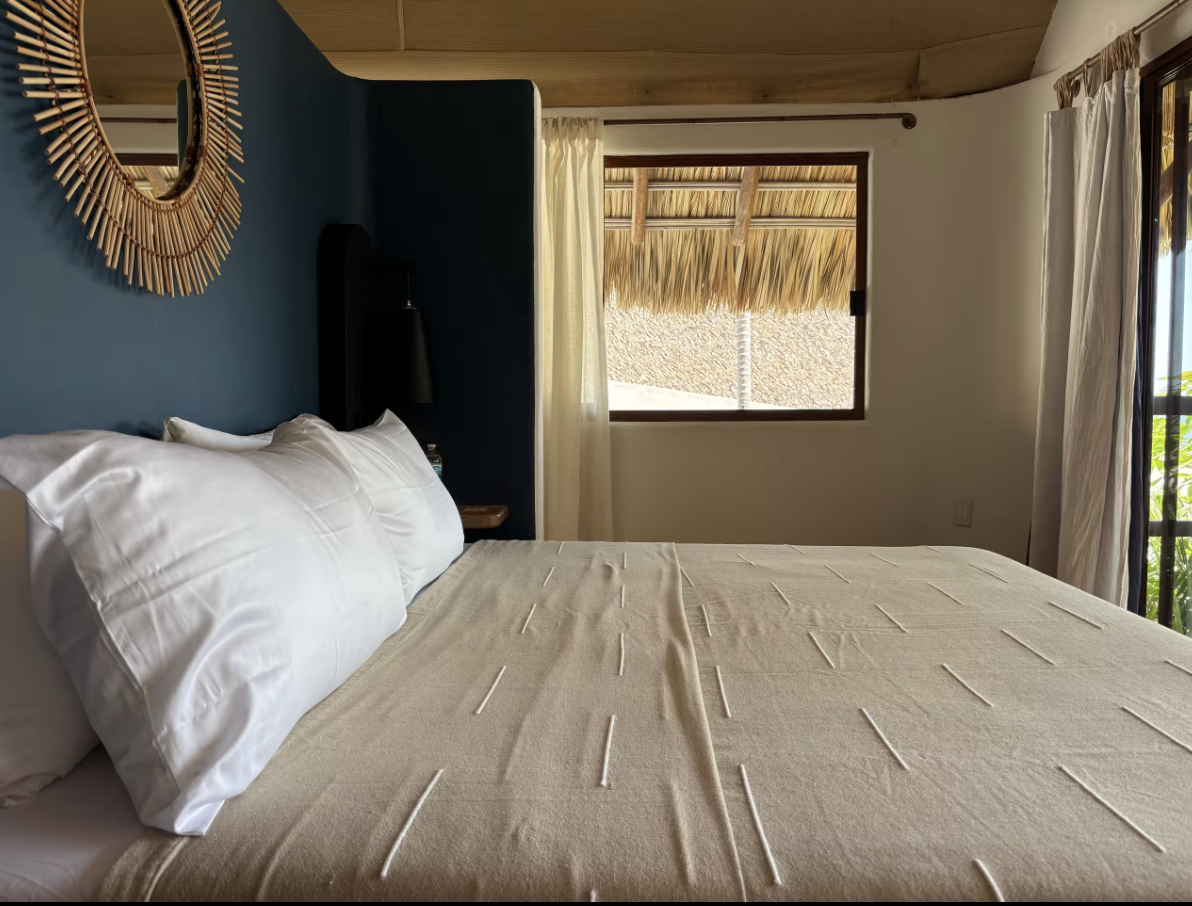 A cozy bedroom with a bed that has white pillows and a beige blanket, a mirror hanging on a blue wall, a window with cream curtains, and a view of a thatched roof outside.