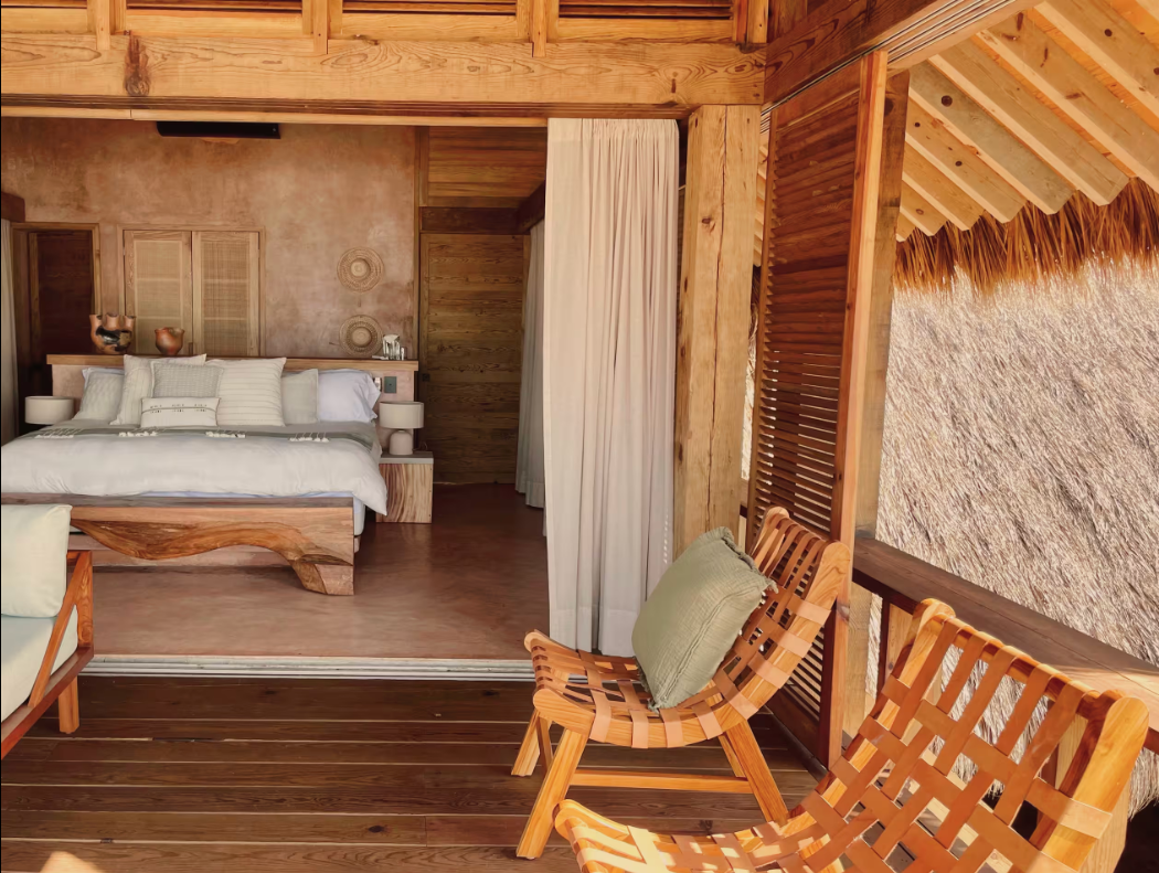 A rustic bedroom with a wooden bed and white bedding, view from a balcony with woven wooden chairs and wicker wall.
